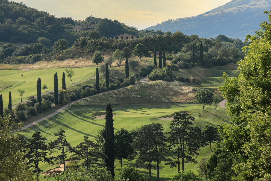 Scenic view of a golf course surrounded by trees on rolling hills with a house in the distance under a partly cloudy sky. Golf Italy. Caddie & Co. | Discover exclusive golf trips.