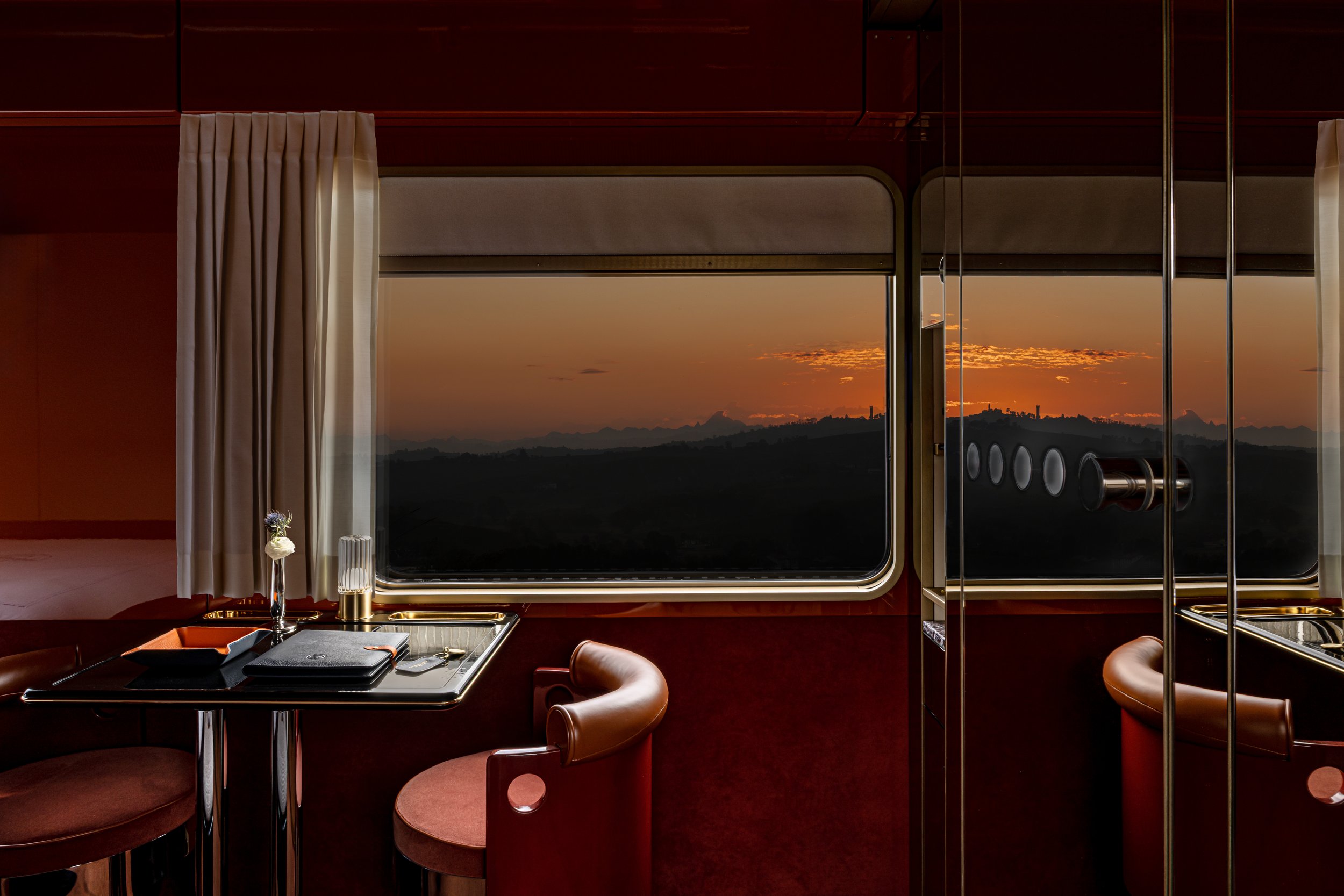 Interior of a luxurious train compartment with a large window showing a sunset over mountains, featuring a small table with a flower, notebook, and glass, and two leather chairs.