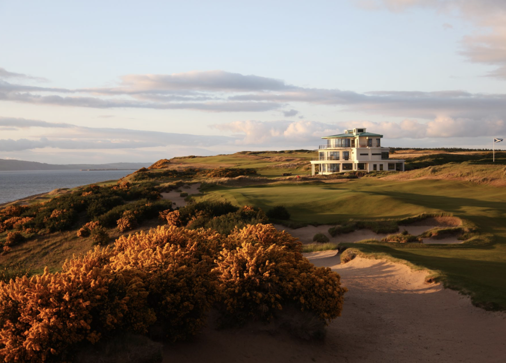 A golf course with vibrant green grass, sand bunkers, nearby shrubs with orange foliage, and a large modern house on a hill overlooking the water, under a partly cloudy sky. Golf Scotland. Cabot Highlands. Caddie & Co. | Discover exclusive golf trips