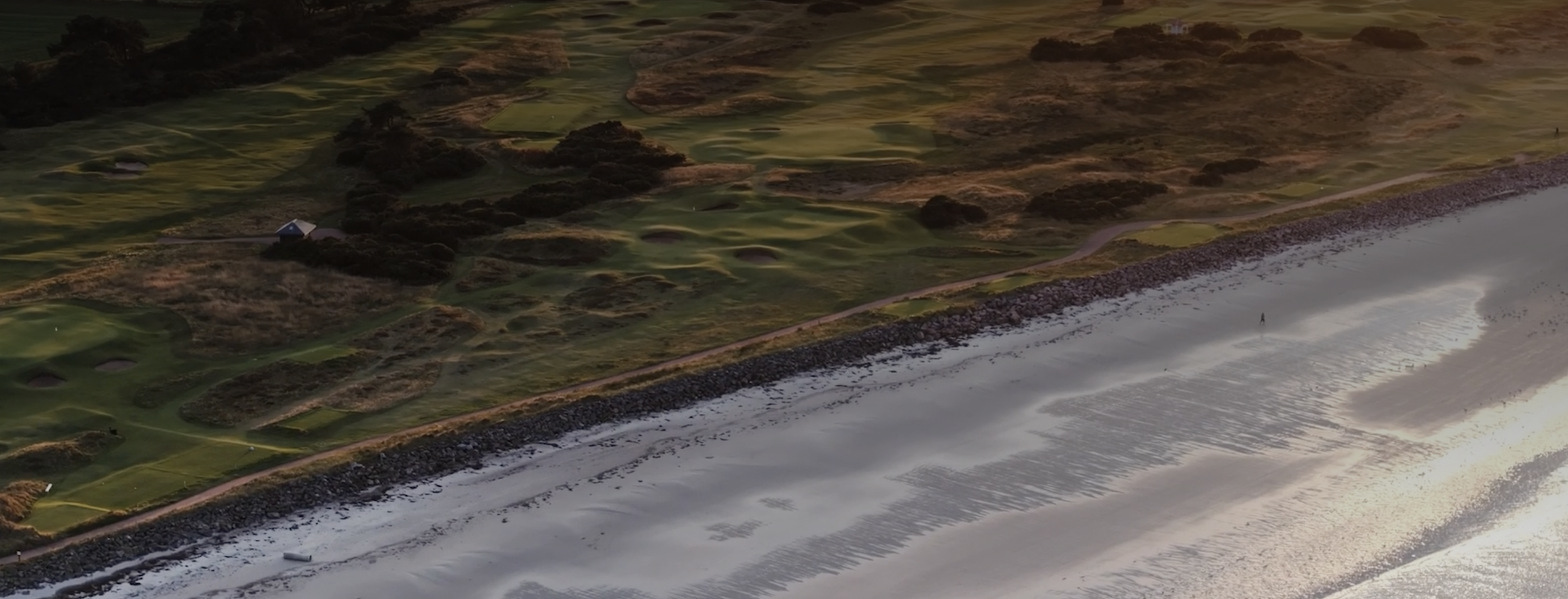 Aerial view of a coastal golf course with green fairways, surrounded by rocky shoreline and sandy beach along the ocean.