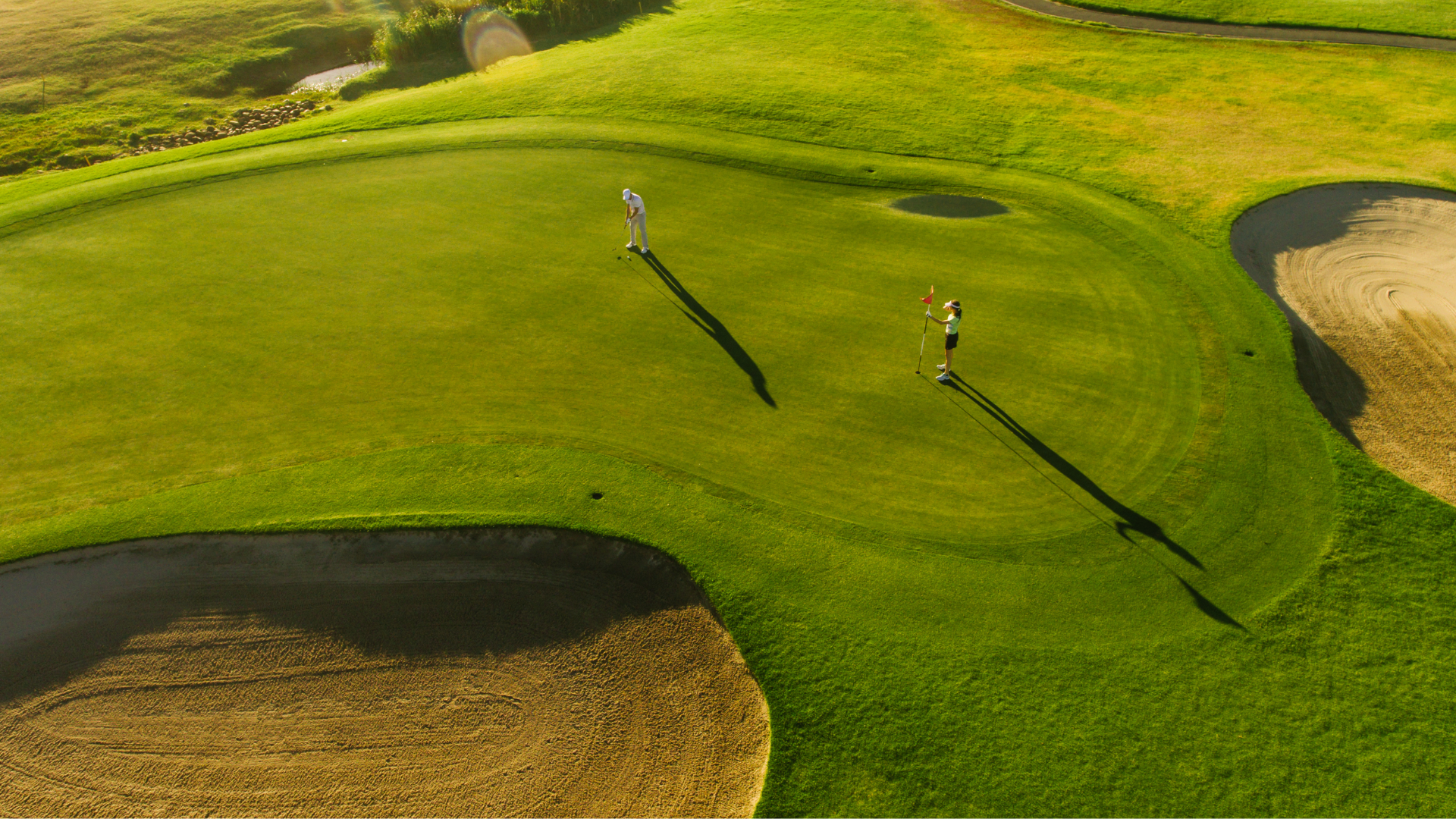 Two golfers on a green golf course, one preparing to putt and the other holding a flagstick, with sand bunkers nearby and a scenic landscape in the background.