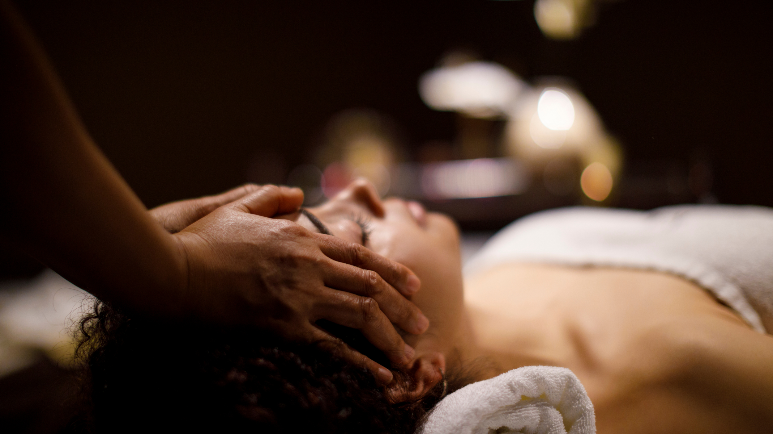 A person receiving a massage with their eyes closed, lying on a massage table in a dimly lit room.