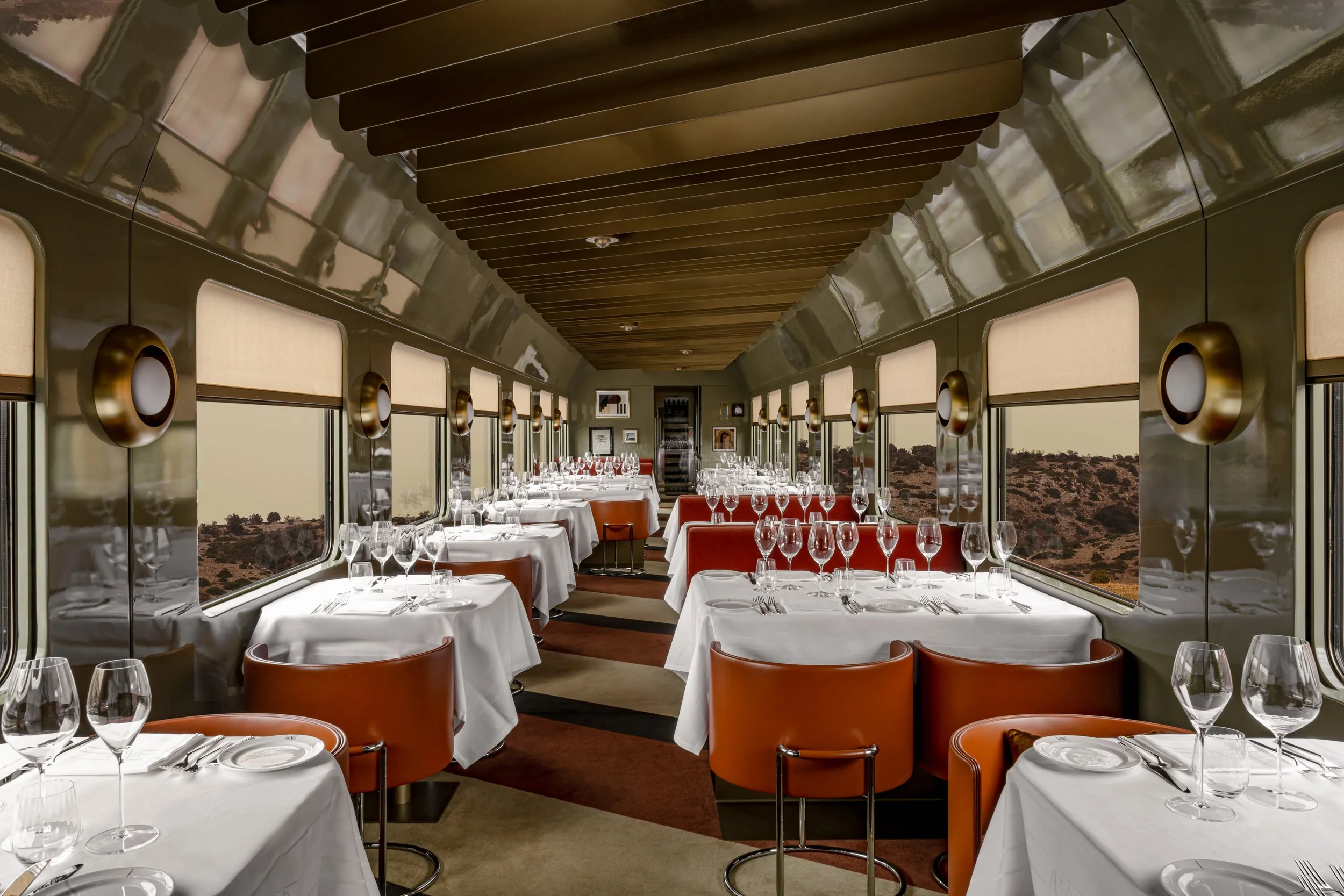 Elegantly set dining car interior with white tablecloths, glassware, and orange chairs, overlooking a desert landscape through large windows.