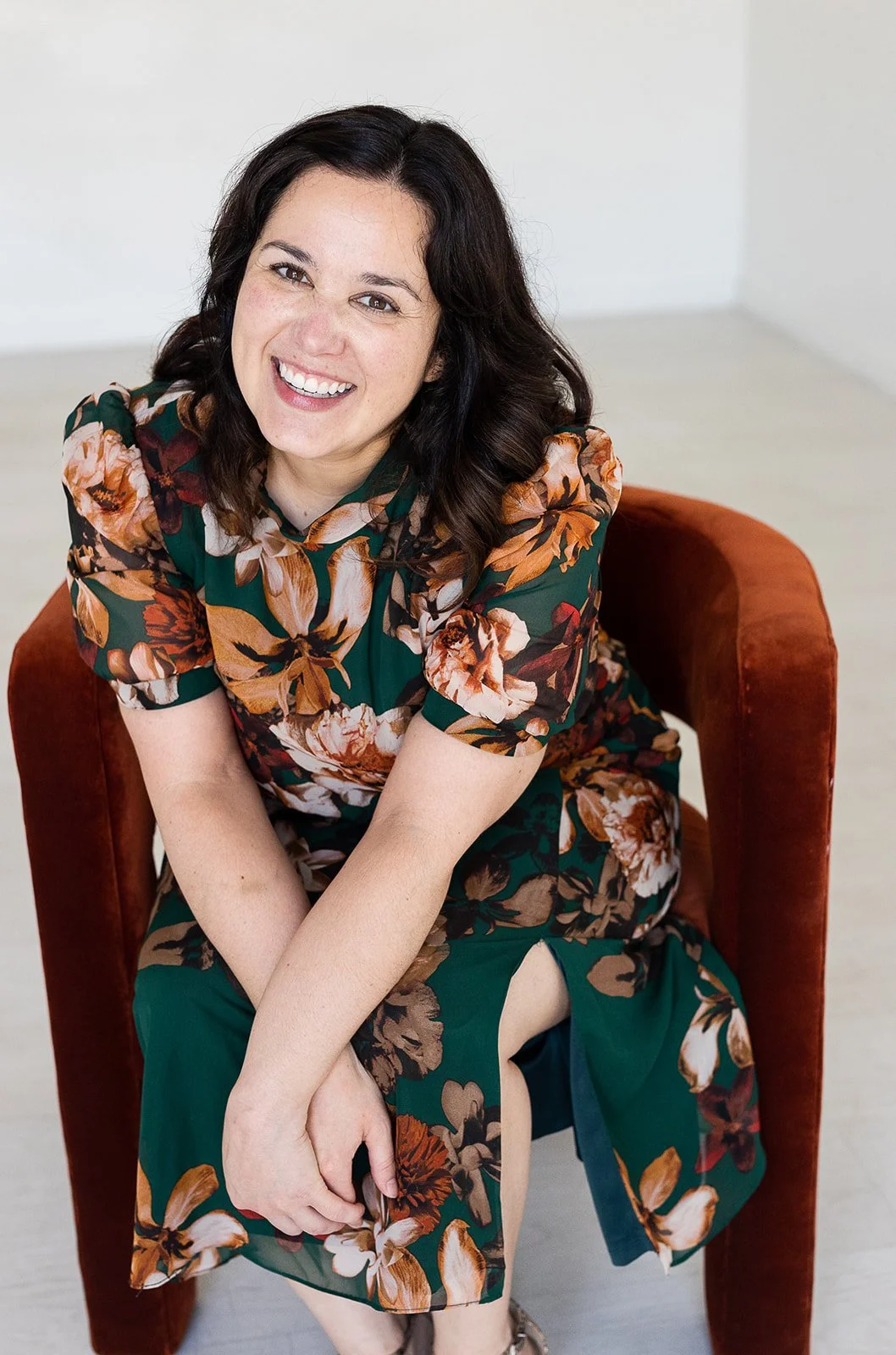 smiling woman with dark hair wearing a green floral dress and seated in a rust colored velvet chair