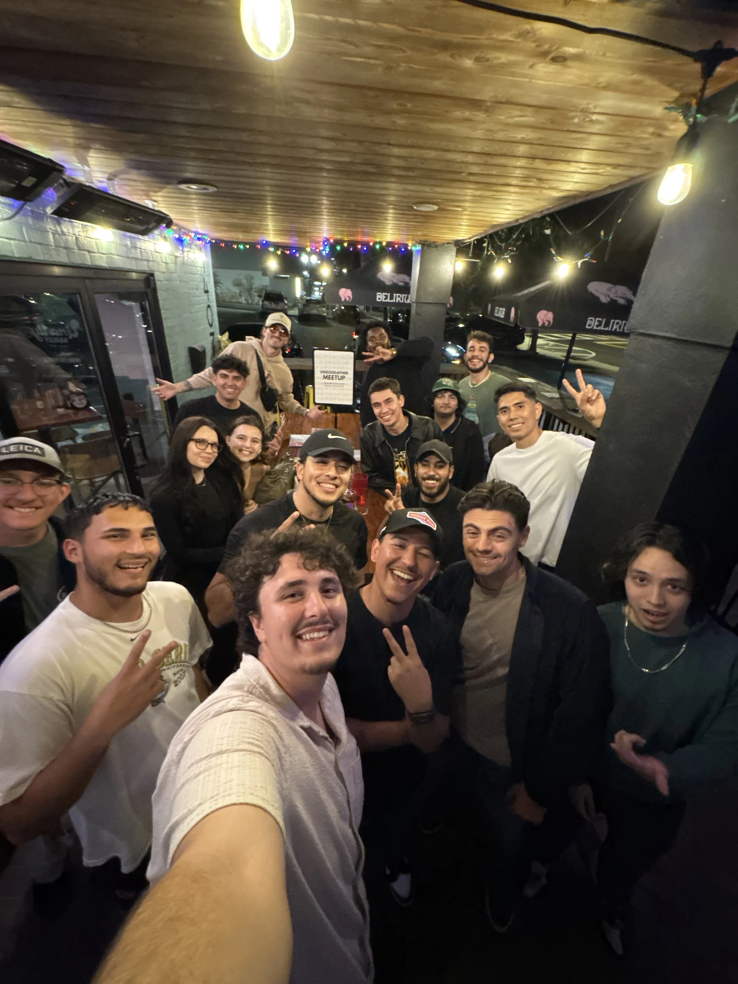 A photo of the Orlando Videographer Meetup showing a group on an outdoor patio area at night, taking a selfie together. The patio has string lights and a wooden ceiling, with parked cars and a building with signage in the background.
