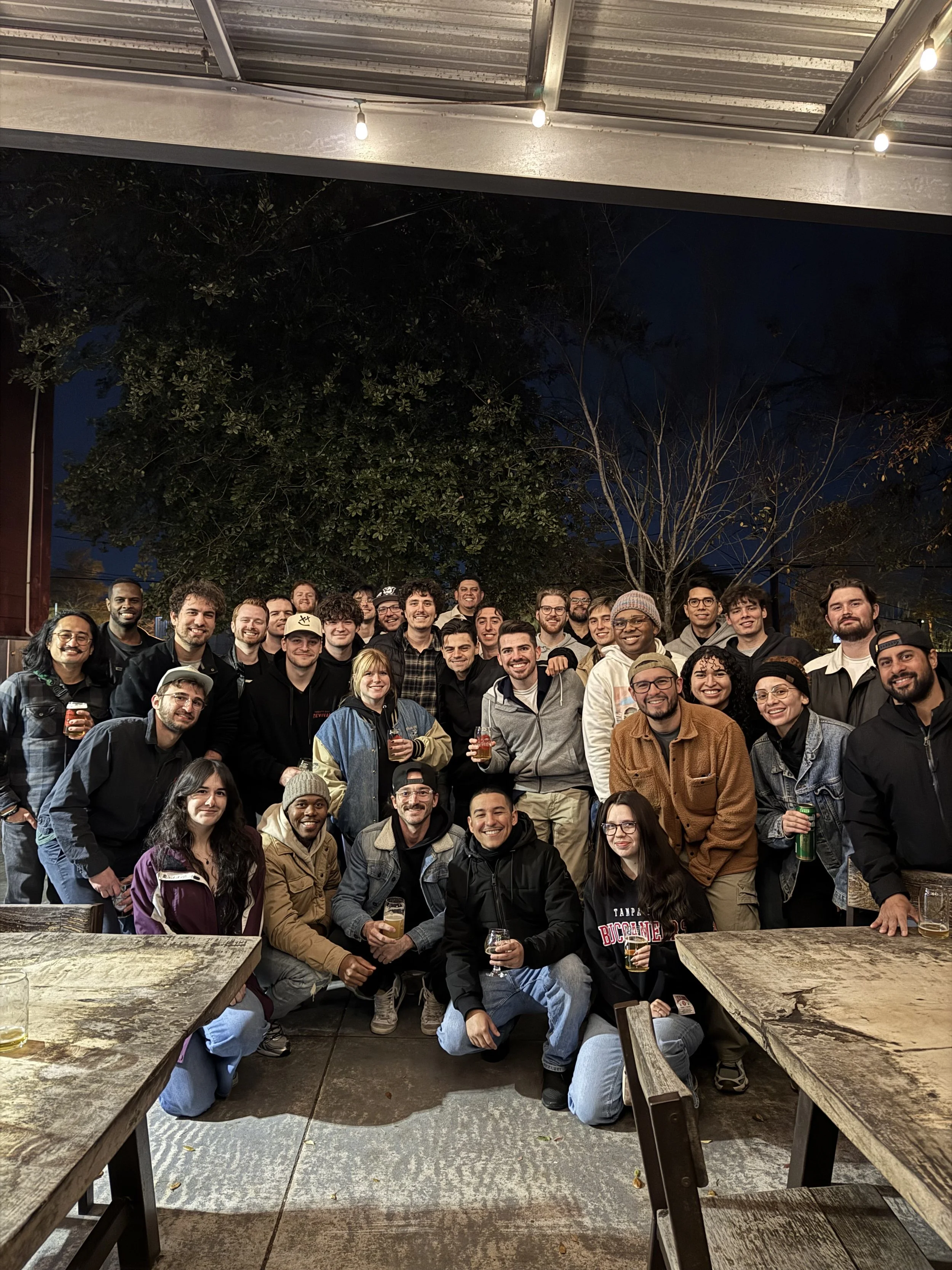 A photo of the Orlando Videographer Meetup showing a large group of people gathered at an outdoor setting during nighttime, smiling, holding drinks, and posing for a group photo beneath a metal roof and string lights with trees in the background.