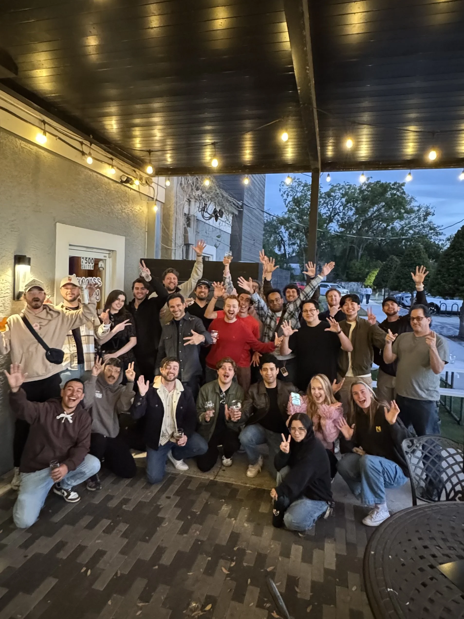 A photo of the Orlando Videographer Meetup showing a large group of people celebrating outdoors in the evening, waving and smiling, with string lights overhead and trees and parked cars in the background.
