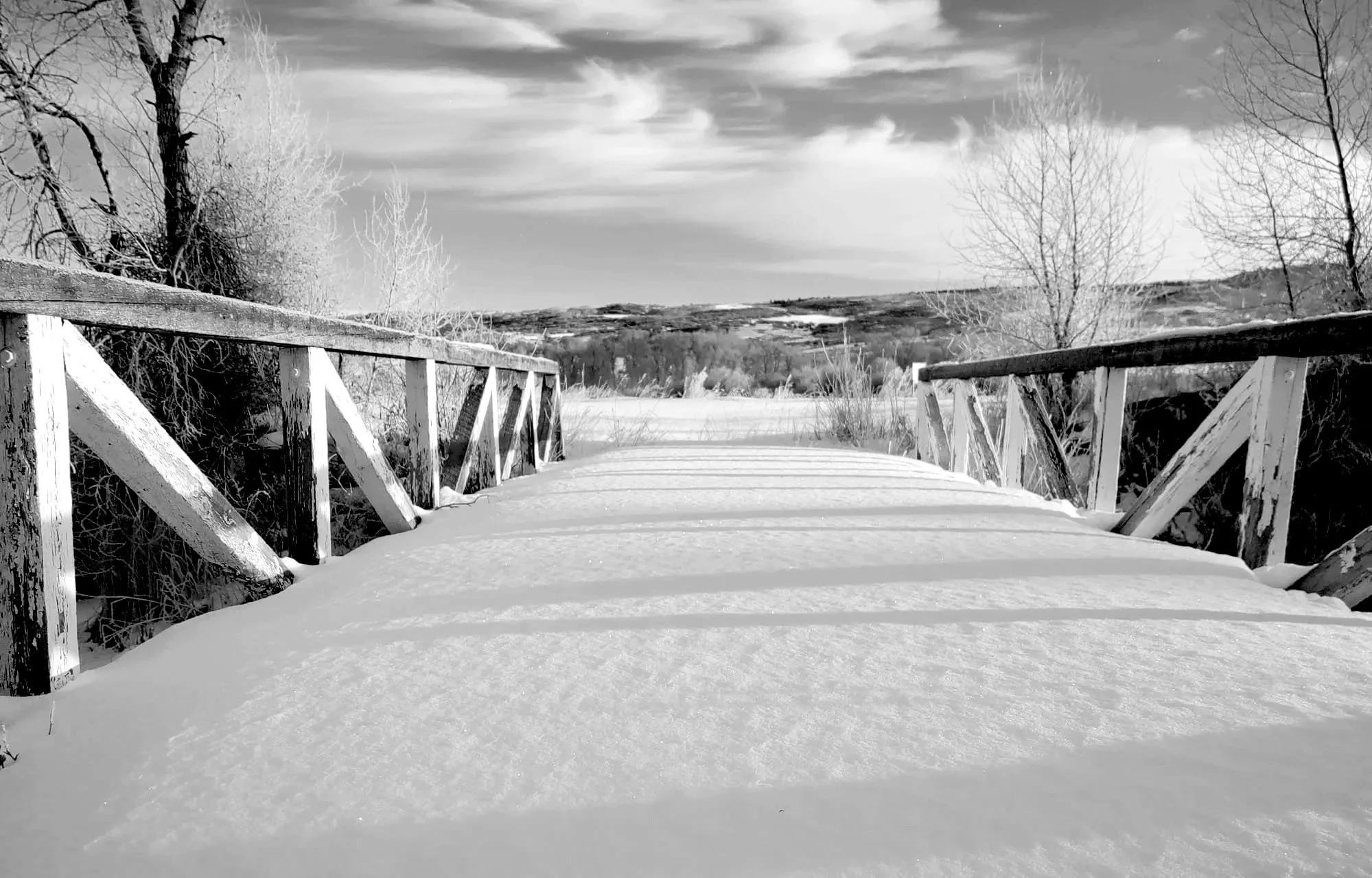 Snow-covered wooden footbridge over a river or stream, with leafless trees on either side and a rural landscape with hills in the background under a cloudy sky.