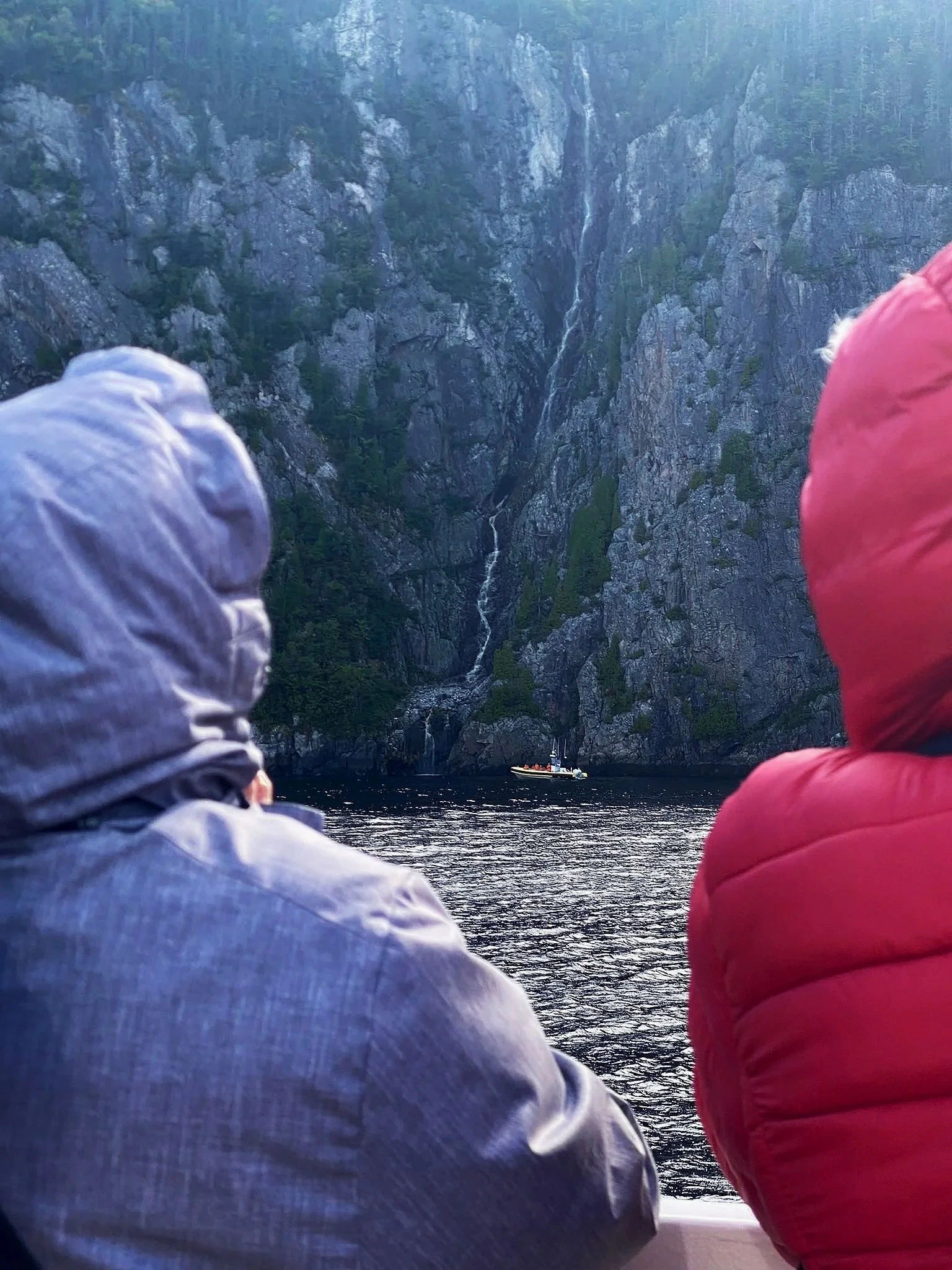 Two people in hooded jackets looking at a waterfall on a tall cliff across a body of water with a boat in the distance.