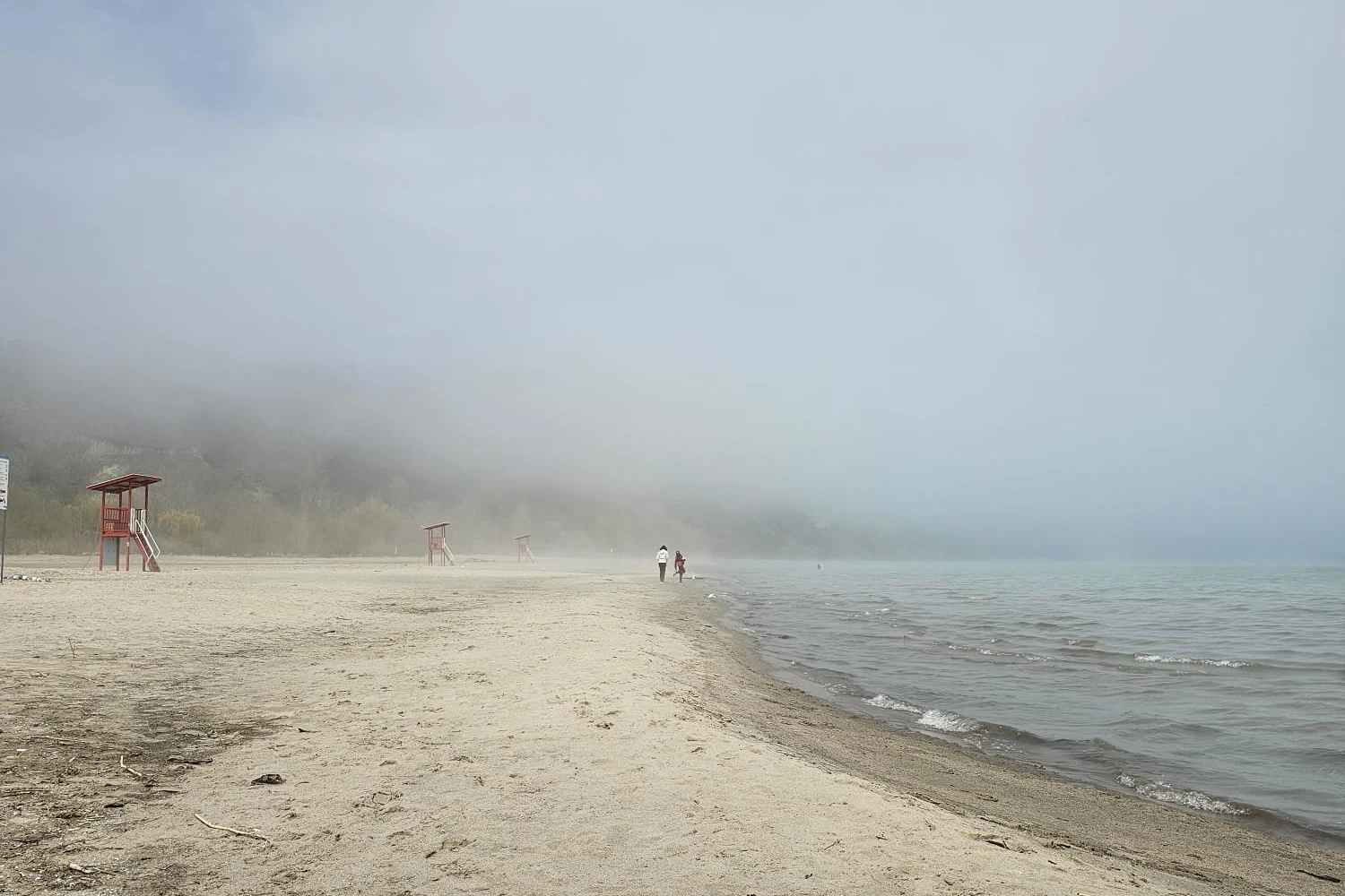 Beach with sand, waves, fog, and lifeguard towers in the distance, with two people walking along the shoreline.