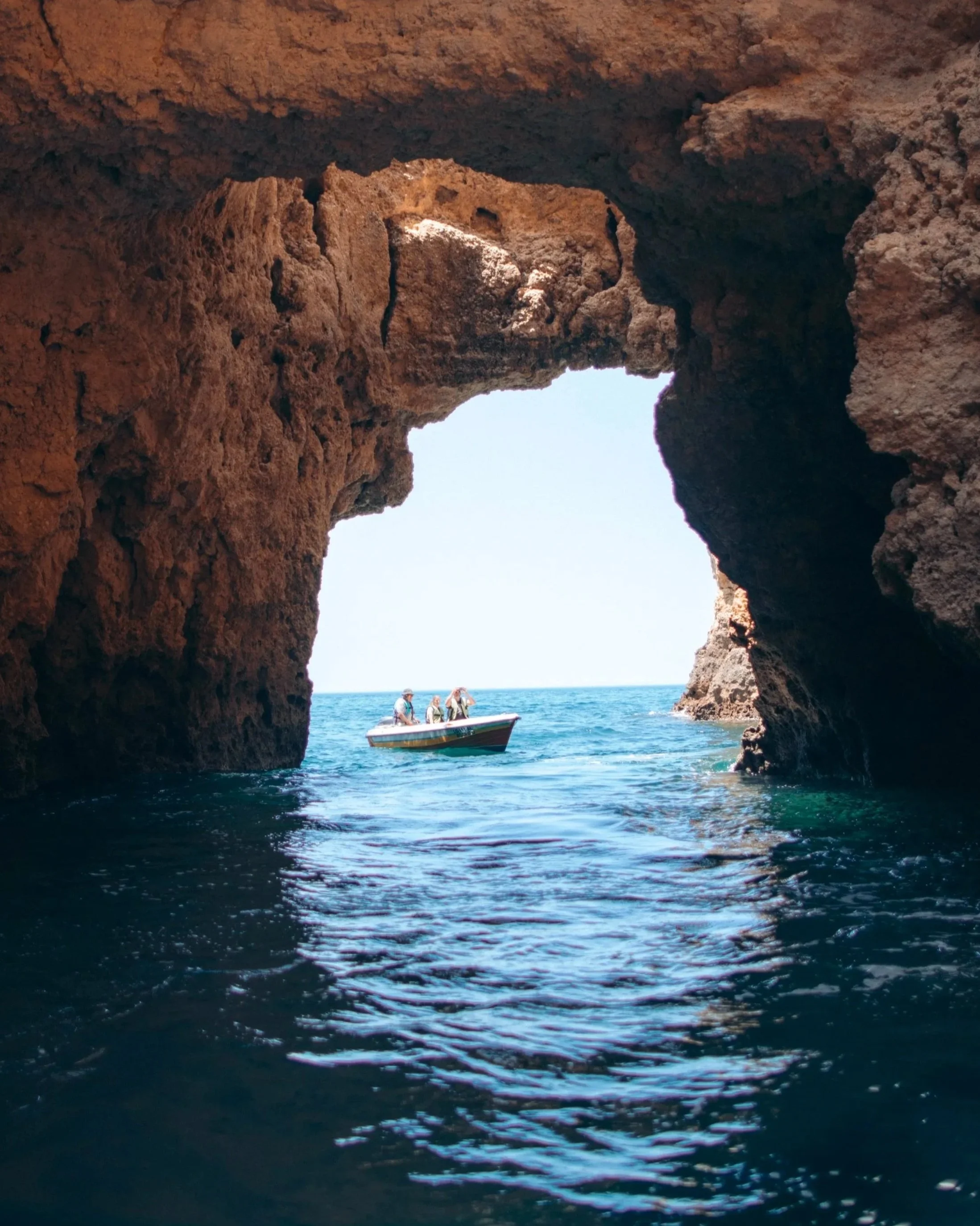 View from inside a rocky sea cave looking out towards a boat with people near the cave entrance and the open ocean beyond.