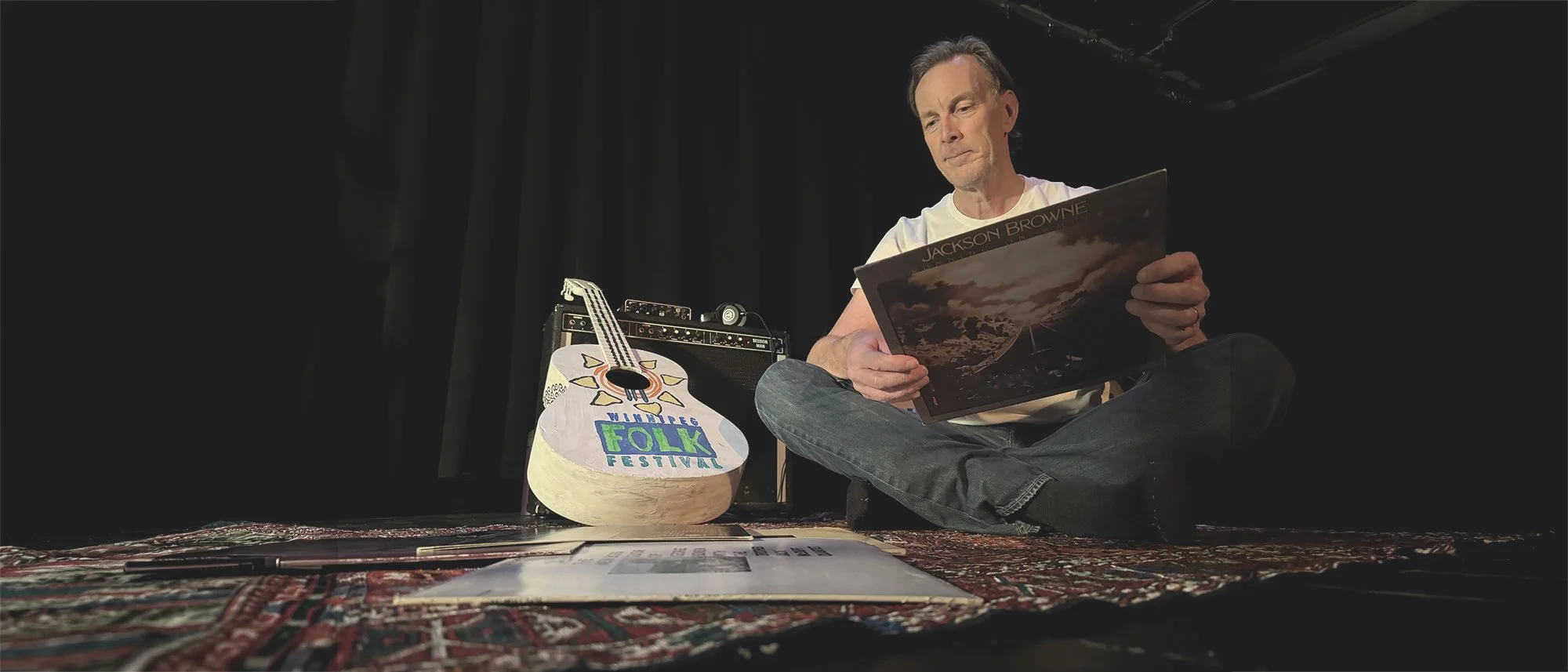 A man sitting on the floor reading a vinyl record album in a dimly lit room. Next to him is a guitar painted with colorful designs and the words 'MINIT FOLK FESTIVAL'. In the background, there is a small amplifier and a black curtain.