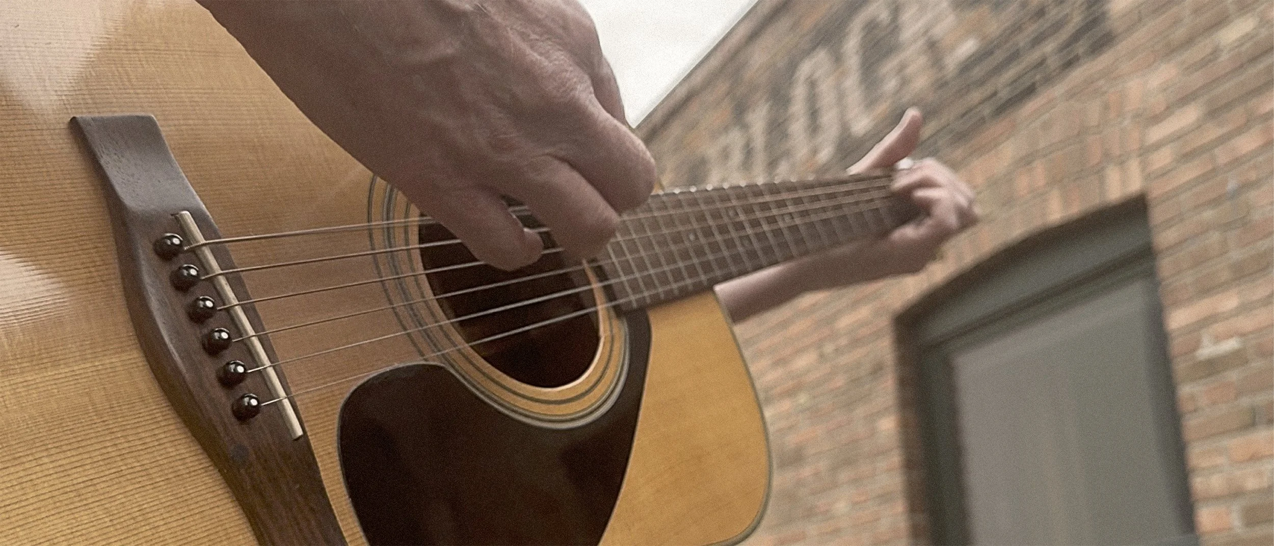 Close-up of a person's hand strumming an acoustic guitar against a brick building background.