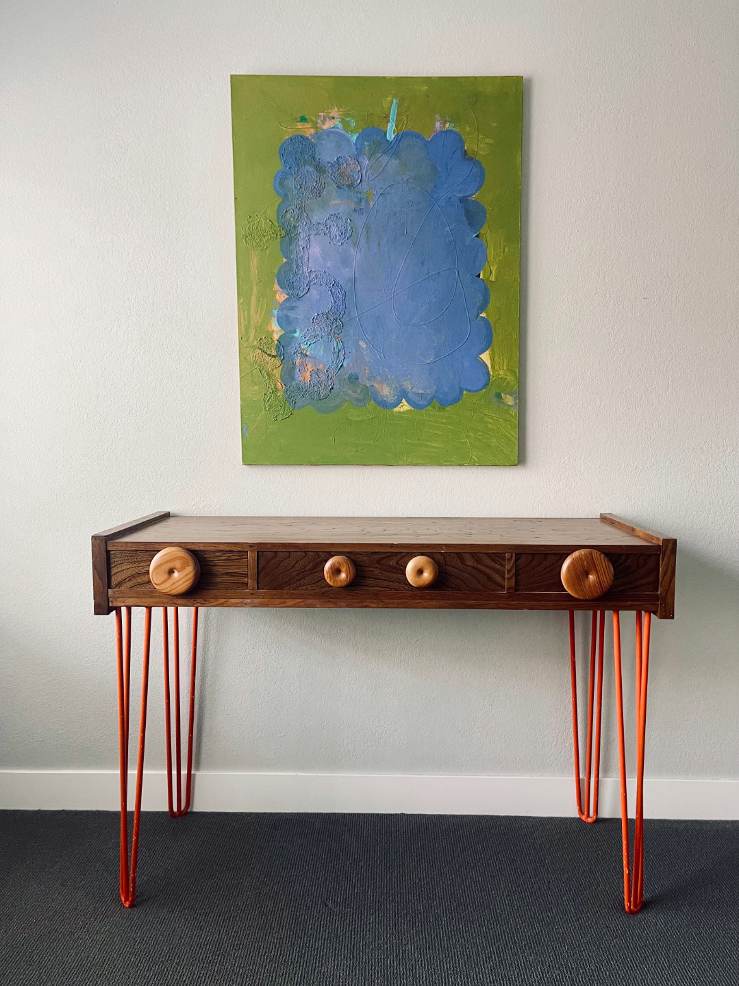 An abstract painting of blue, purple, and pink shapes on a green background, hanging above a wooden table with orange metal hairpin legs and four wooden knobs.