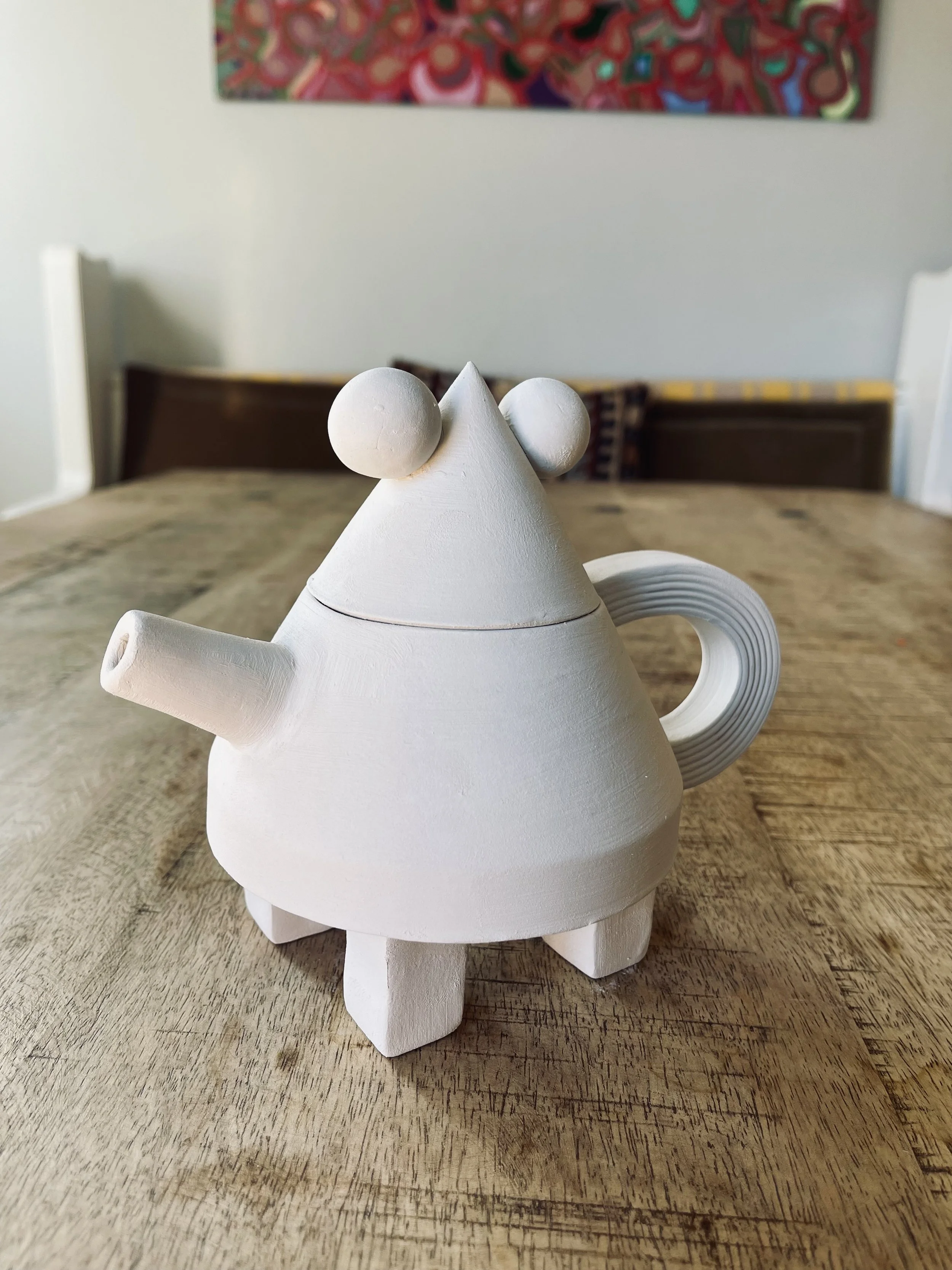 A white ceramic teapot shaped like a cartoon animal with a rounded body, small ears on top, a spout, a handle, and four short legs, sitting on a wooden table.