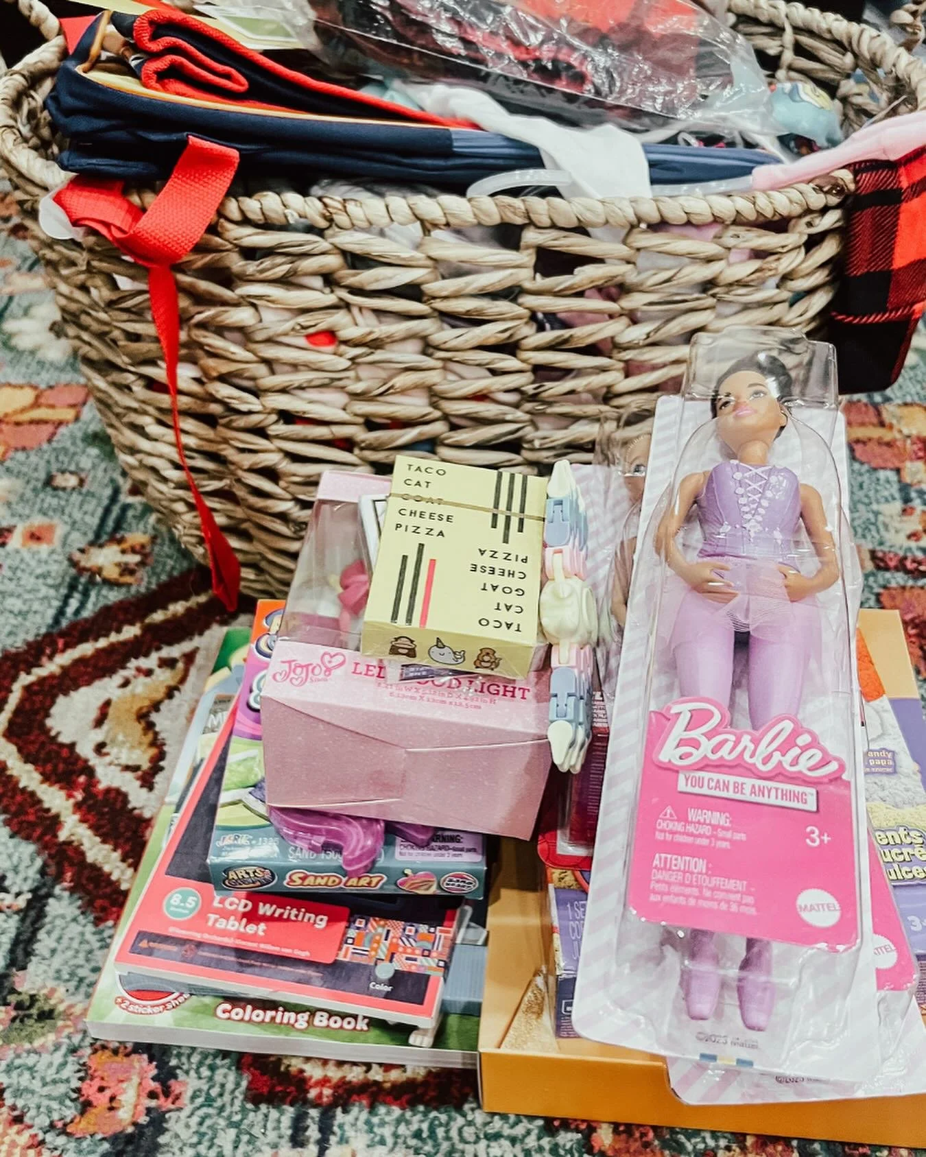 A basket filled with various toys and items, including a Barbie doll in packaging, a pink box with cartoon animals, a coloring book, a tablet for coloring, and a small card with a menu, all placed on a patterned carpet.