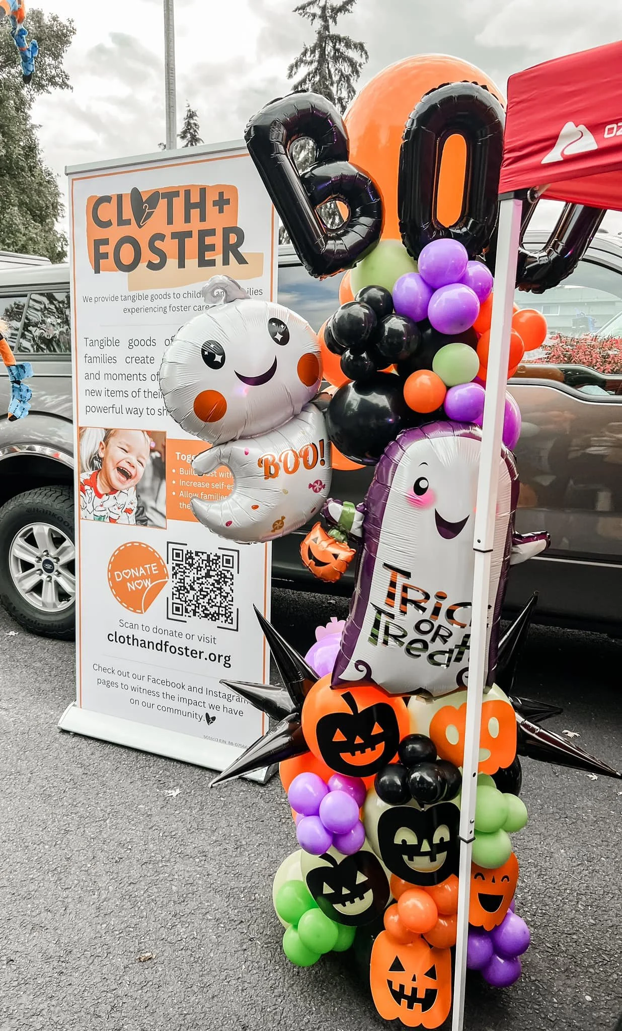 Halloween-themed balloon decorations with pumpkins, a ghost with a "BOO!" message, and a large purple balloon with a smiling face. A sign for Cloth+Foster organization is nearby, promoting donations for foster children, with info, a QR code, and soci