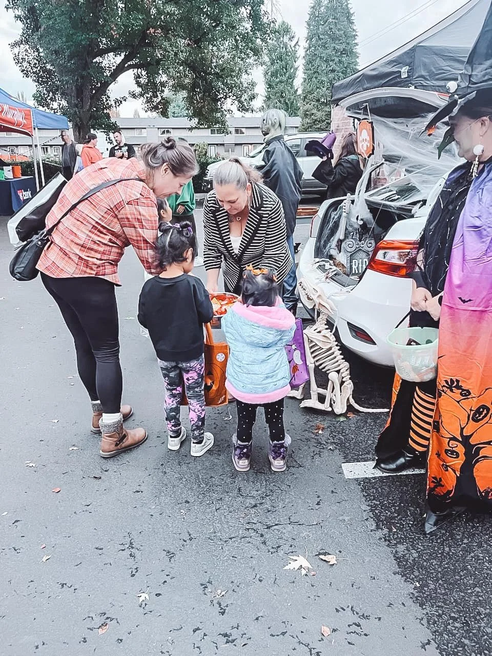 People trick-or-treating at a Halloween event, including children in costumes and adults, with a car decorated for Halloween in a parking lot.
