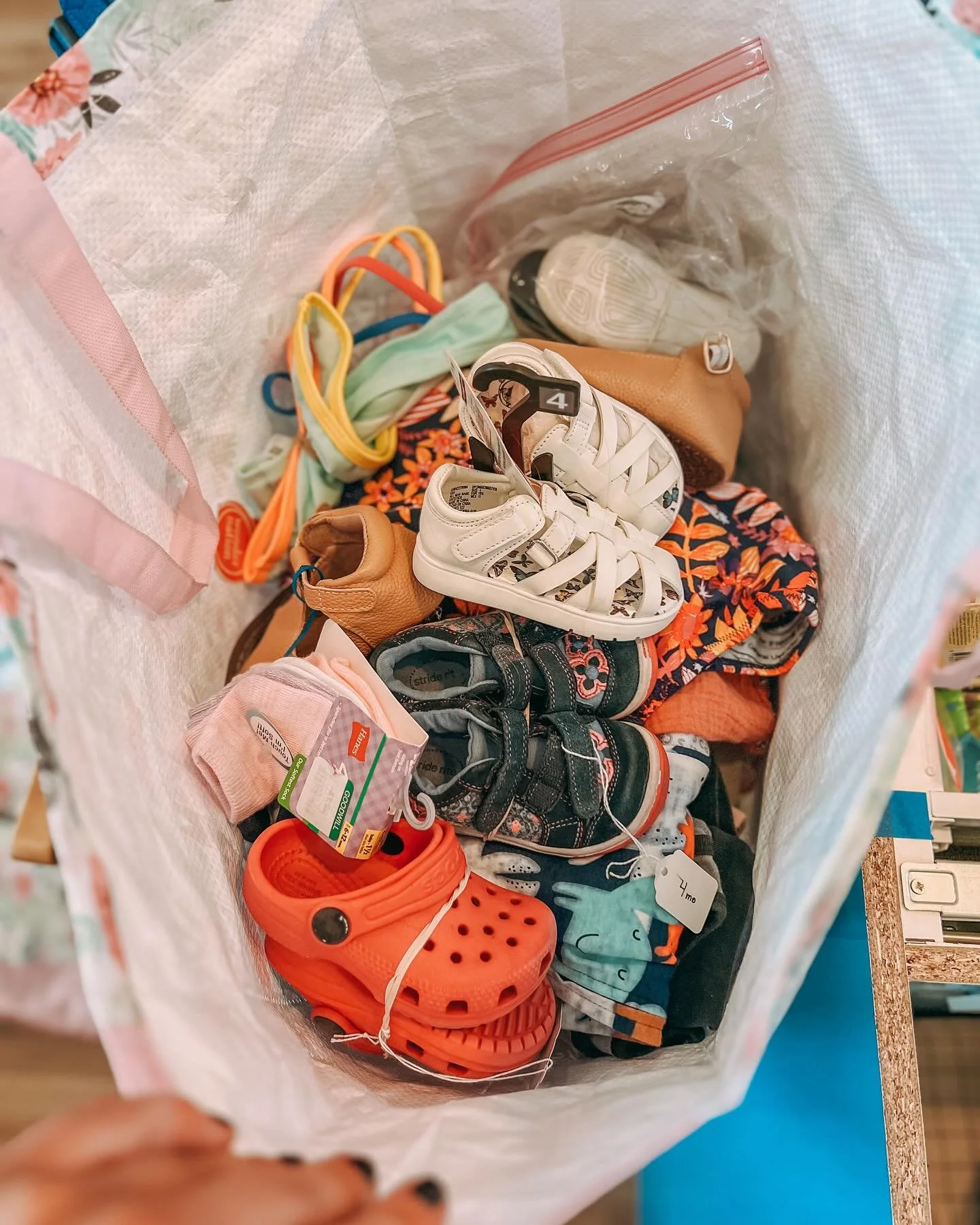 A large shopping bag filled with children's shoes, socks, and sandals, including a pair of red Crocs, white sandals, and various sneakers and boots, with some miscellaneous clothing items visible at the bottom.