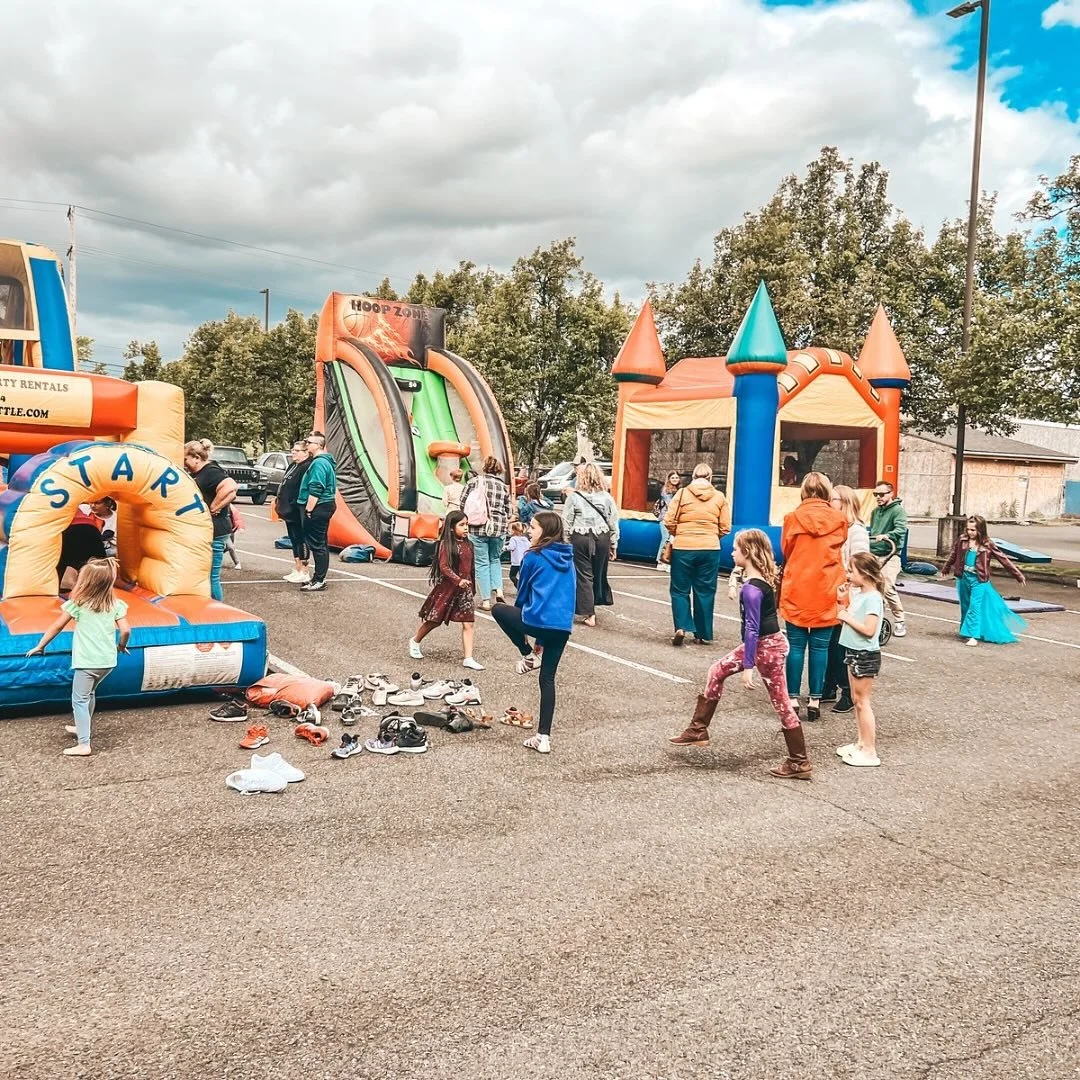 Children and adults at a bounce house party in a parking lot, with various inflatable slides and castles, shoes scattered on the ground, and cloudy sky overhead.