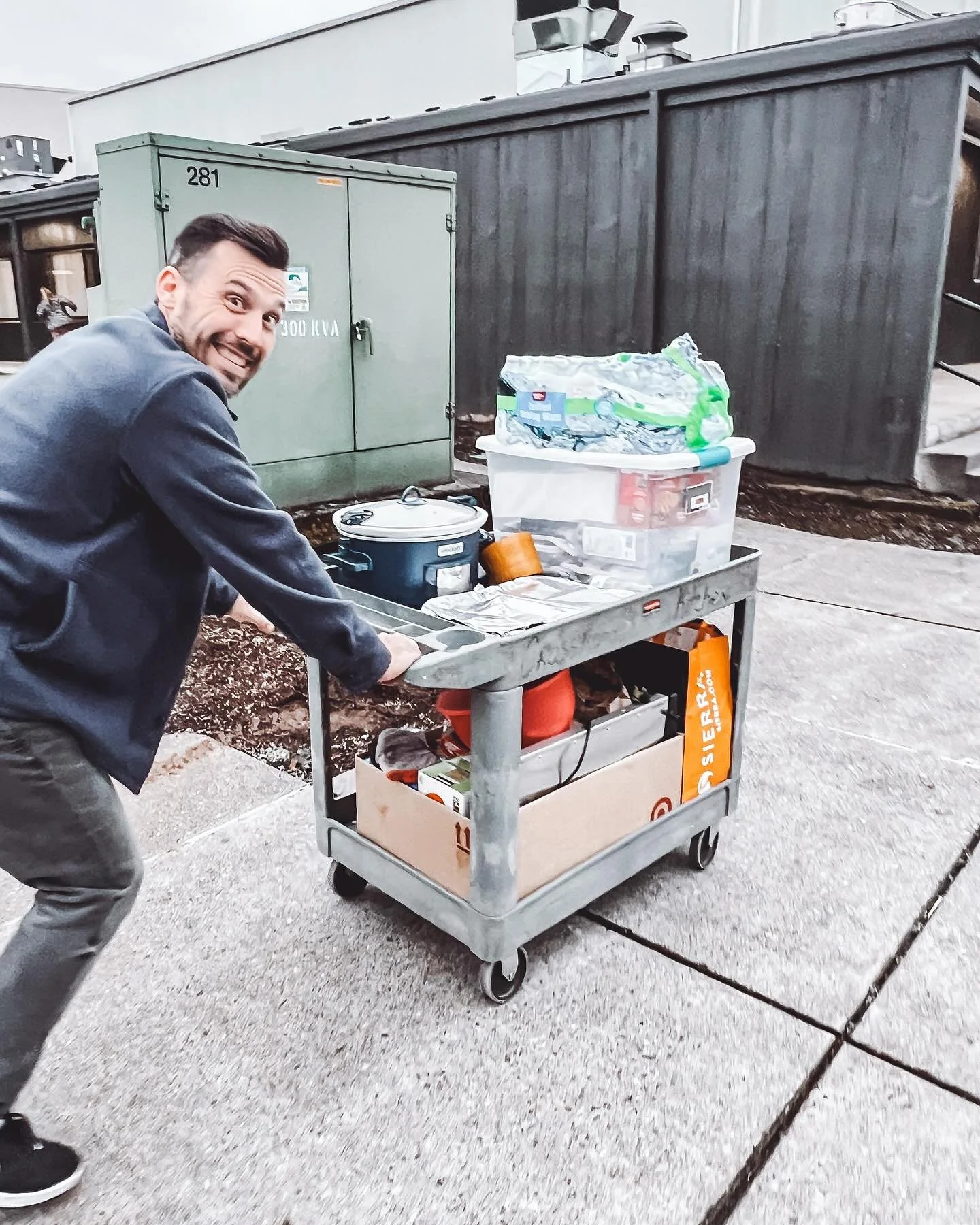 A man smiling and pushing a metal cart filled with boxes and containers headed to a support group for caregivers. 