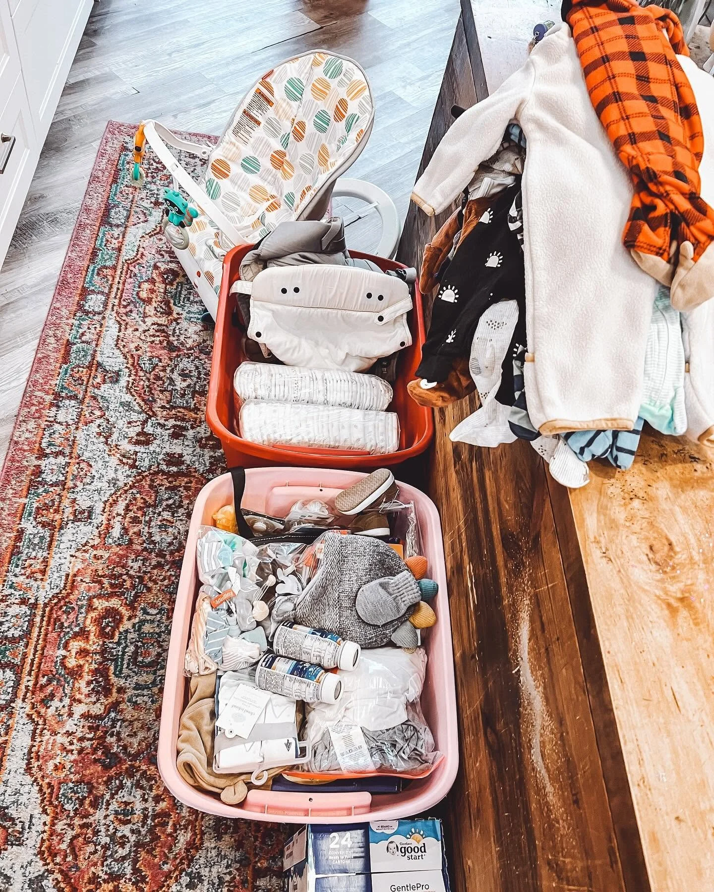 Two plastic storage bins filled with baby supplies, next to a high chair, on a rug and wooden floor.