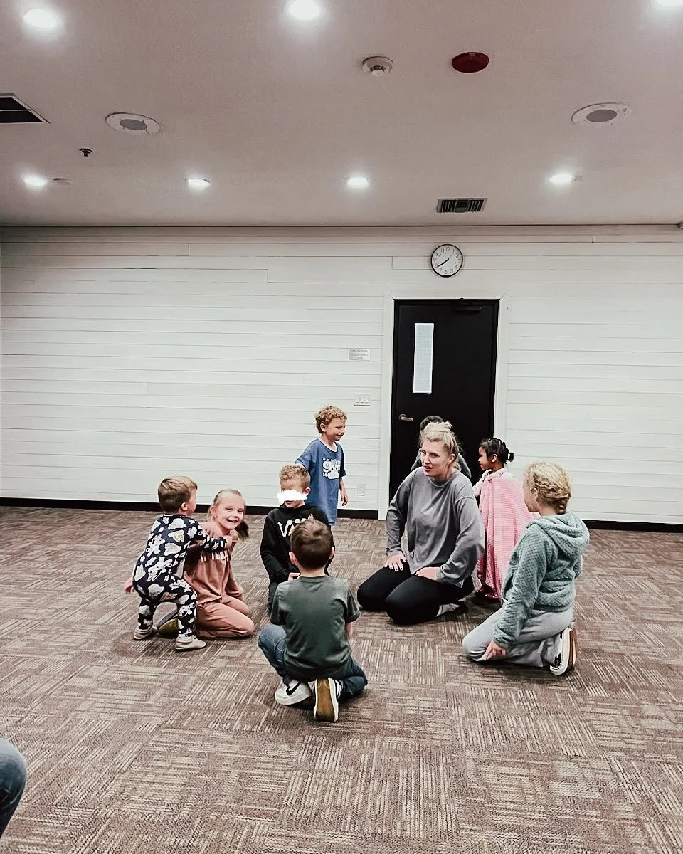 A woman, sitting on the floor, interacts with a group of children in an indoor room with white walls, brown carpet, and a black door. The children are kneeling or sitting around her, some smiling and engaged.