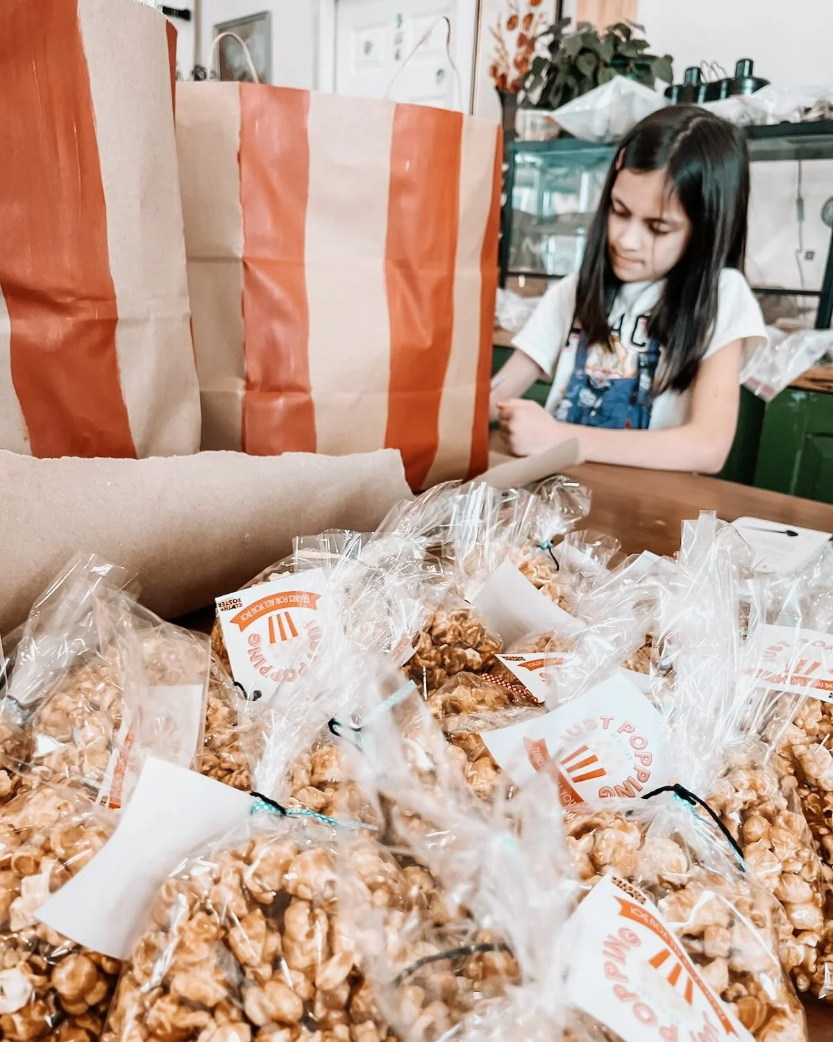 A young girl with long dark hair wearing a white shirt and denim apron behind a table with caramel popcorn bags in plastic packaging and a large paper bag in the background, in a kitchen setting.