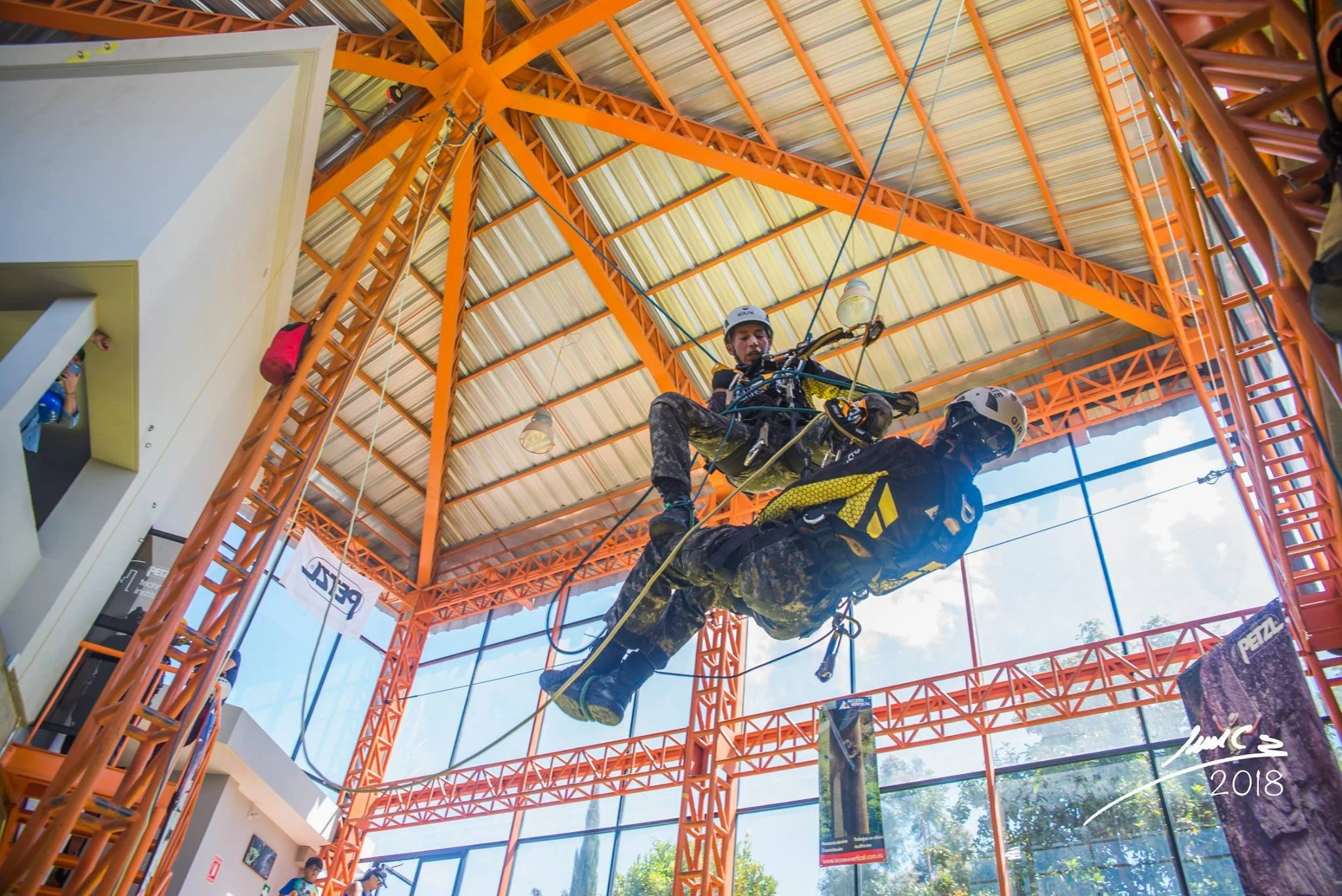 Dos personas practicando puenting en un interior con estructura de metal naranja y techos altos de lámina, con uno de ellos en el aire conectado a cuerdas de seguridad.