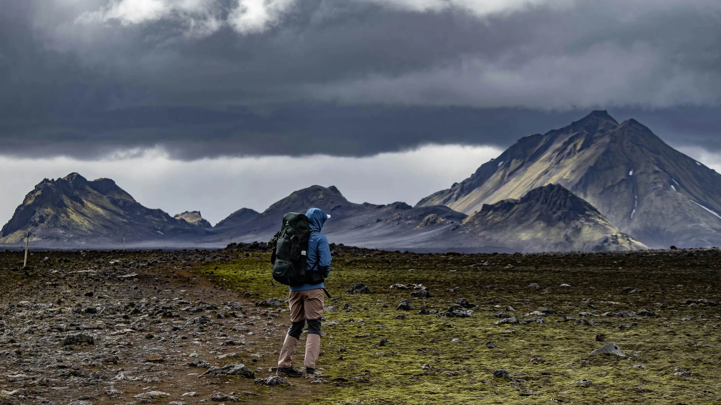 Persona con mochila de senderismo caminando por un paisaje montañoso con cielo nublado.
