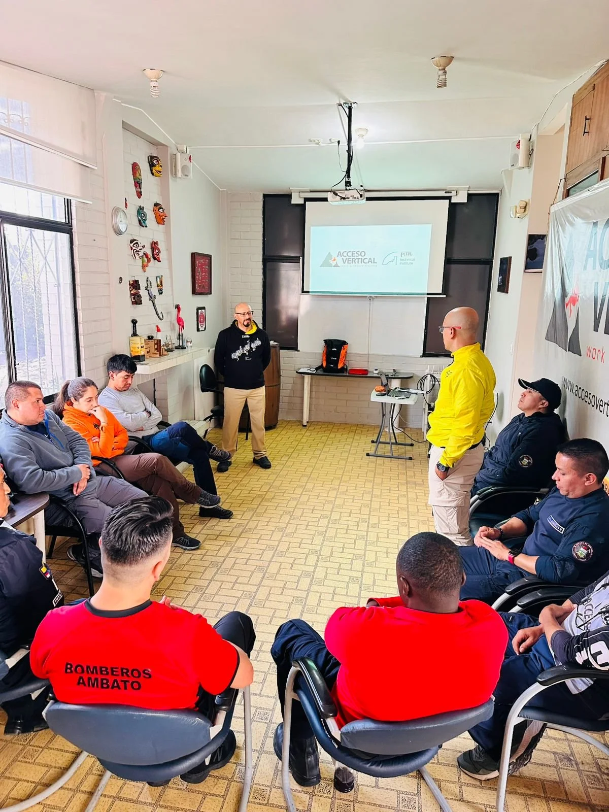 Reunión de entrenamiento o capacitación con varias personas en un aula, algunos uniformados y otros civiles, en una capacitación sobre acceso vertical, en una sala con pantalla y decoraciones en la pared.