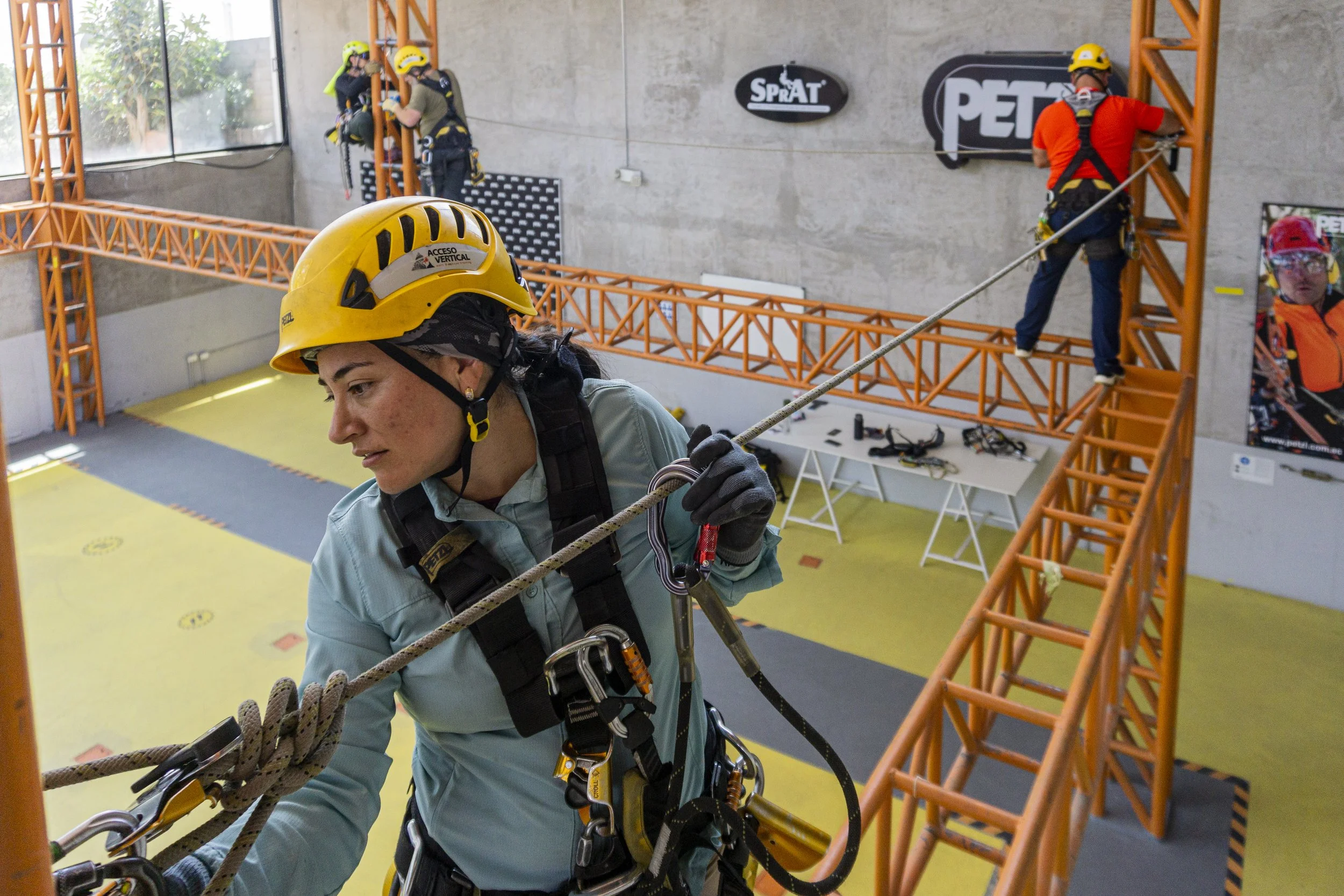 Personas con cascos y arneses en entrenamiento de escalada en interior, con paredes de escalada y estructuras de color naranja.