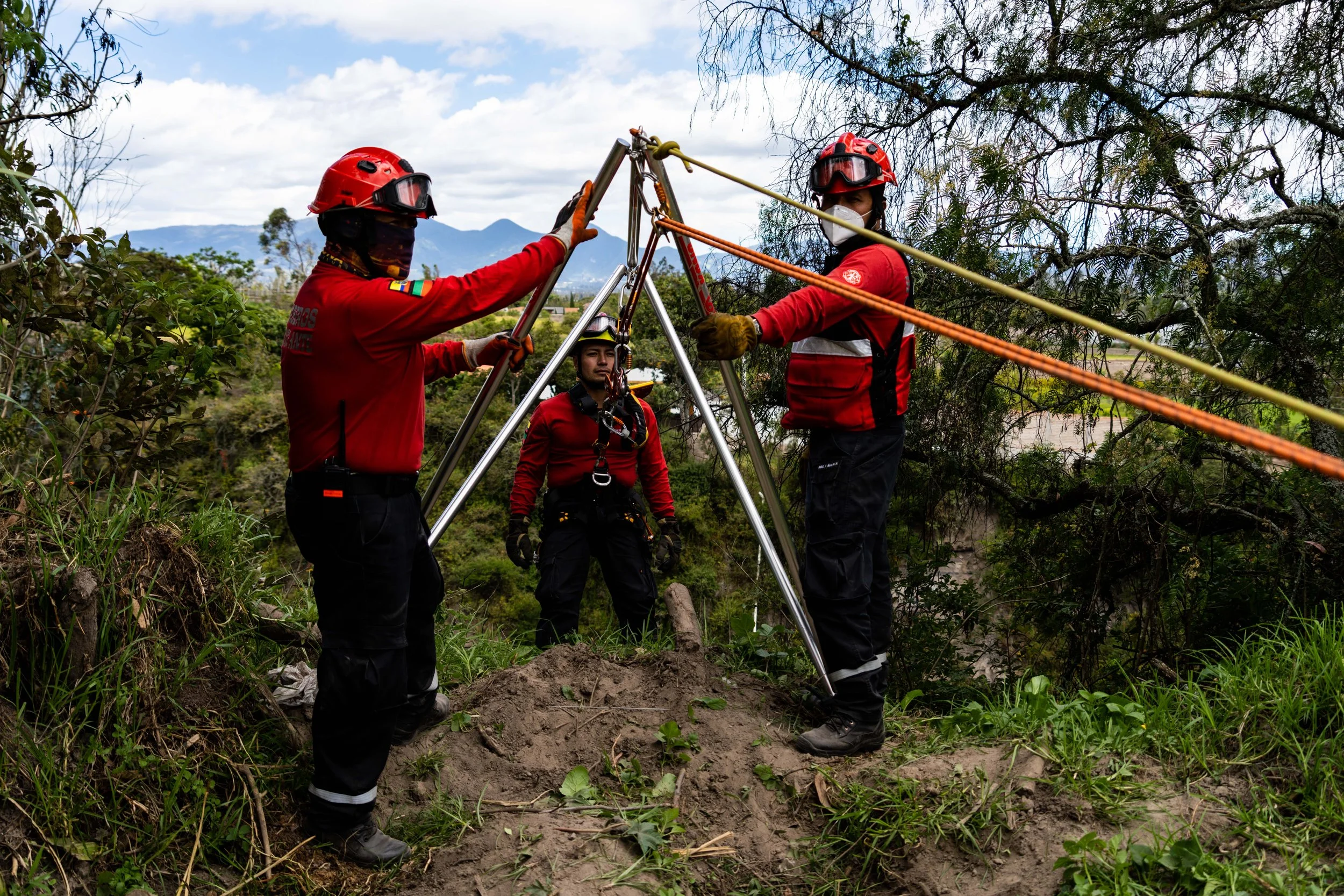 Tres rescate en montañas, dos con cascos rojos y uno con casco amarillo, usando arneses y cuerdas para estabilizarse sobre terreno de tierra y vegetación.