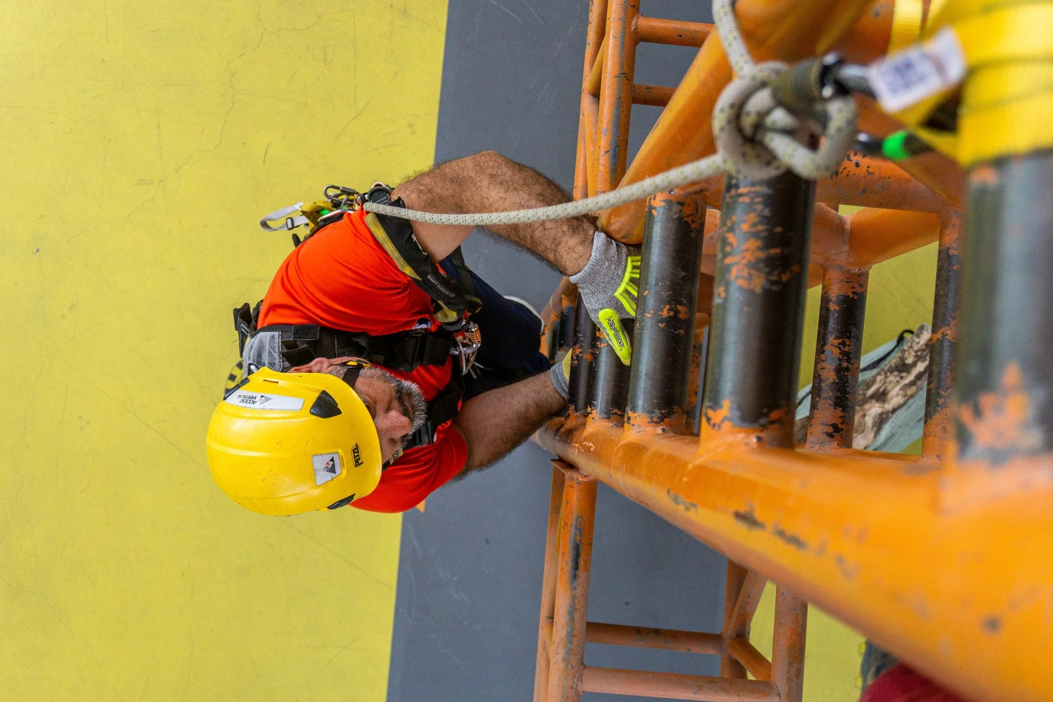 Un hombre usando casco amarillo, camiseta roja y arnés de seguridad está en una estructura de escalada, sosteniéndose con las manos mientras cuida las medidas de seguridad.