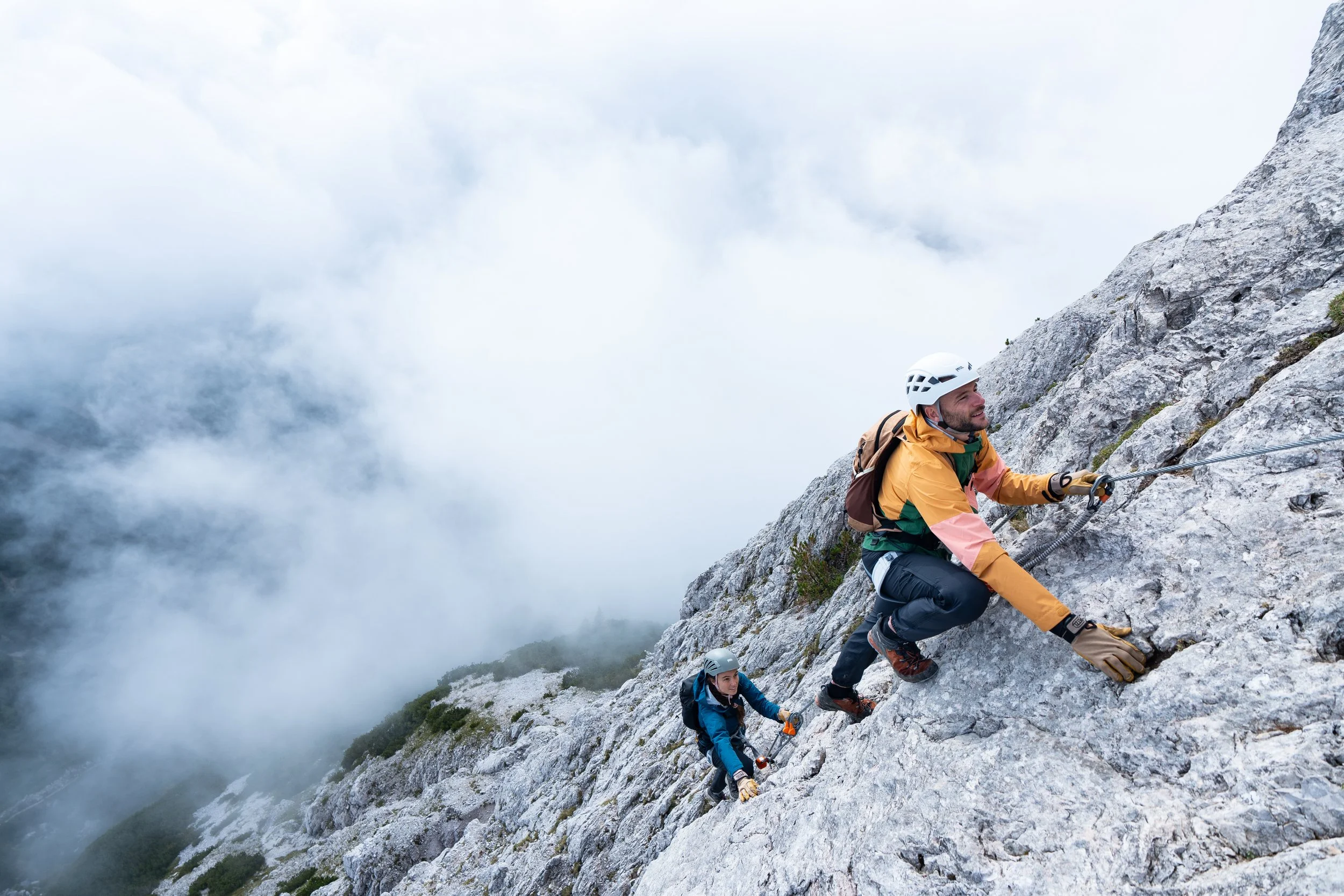 Dos escaladores subiendo una montaña rocosa con nubes y cielo gris de fondo.