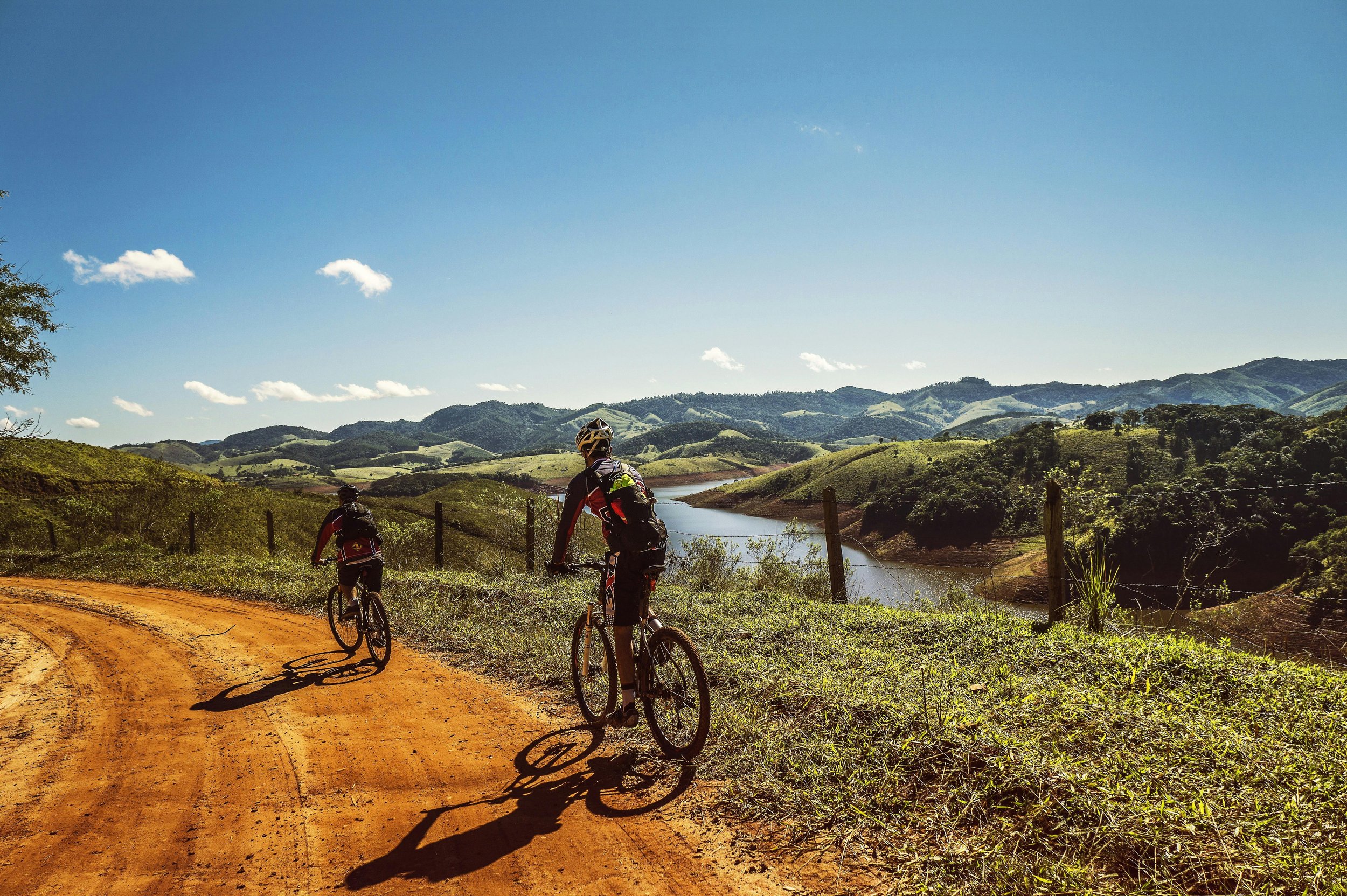 Dos personas en bicicleta sobre un camino de tierra en un paisaje montañoso con río y césped verde bajo un cielo despejado.