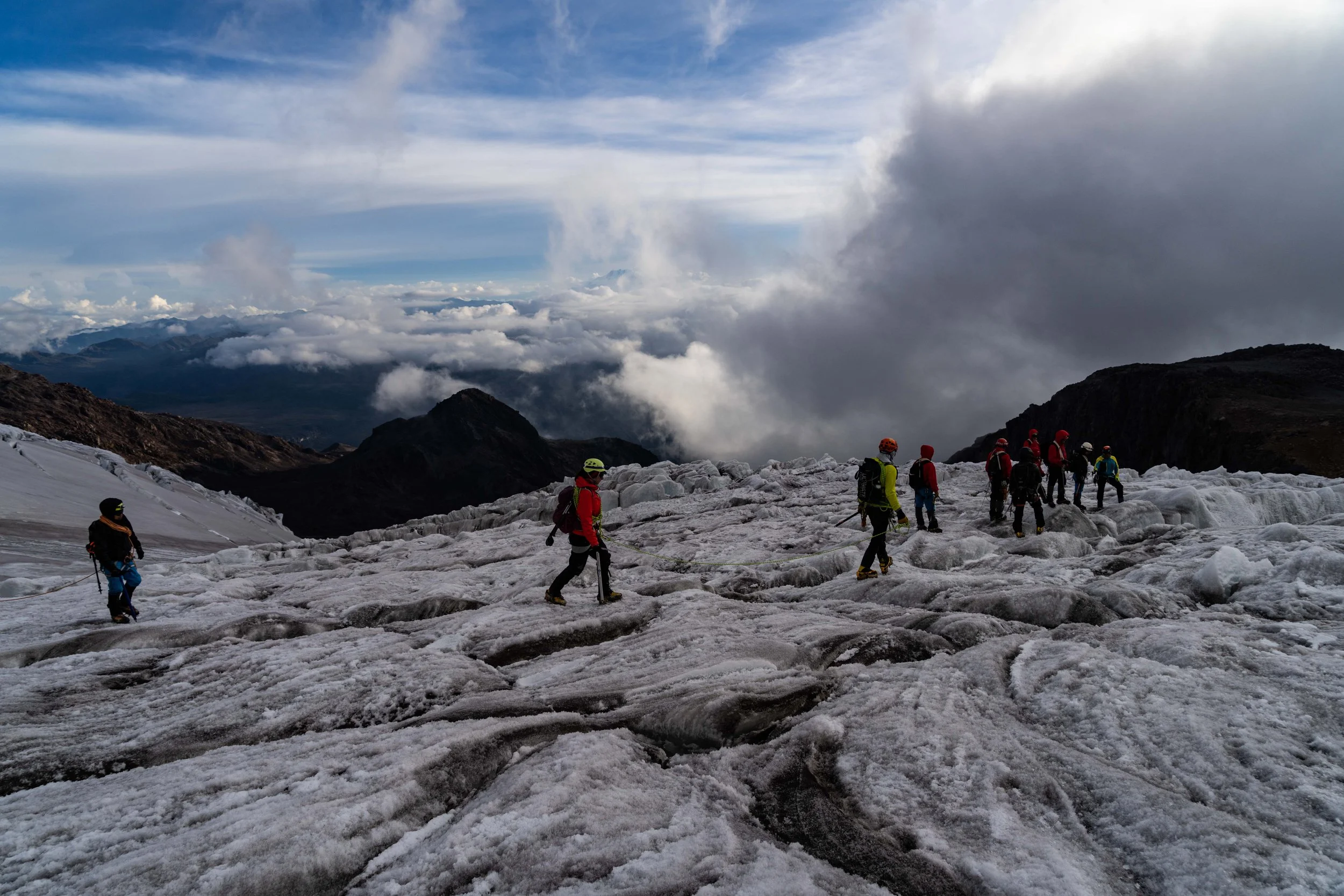 Grupo de montañistas en una nieve glaciar en una cordillera con nubes y cielo parcialmente despejado.