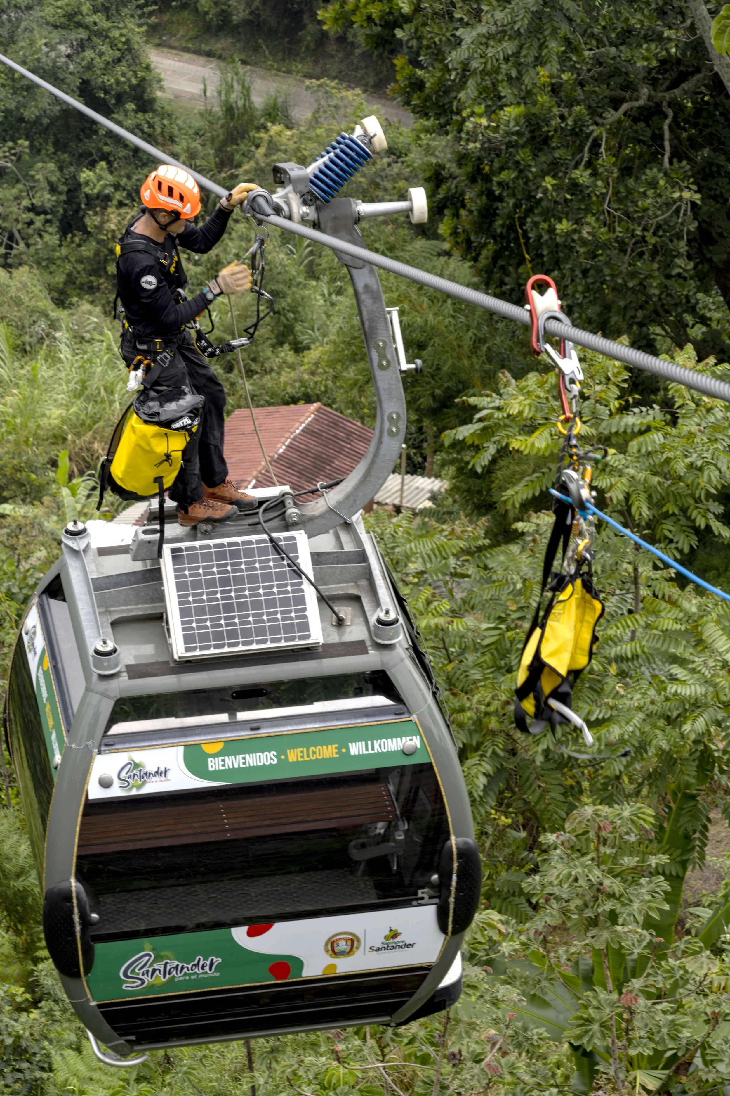Un trabajador, equipado con casco y arnés, ajusta el cable de un teleférico en medio de un entorno con árboles y vegetación.