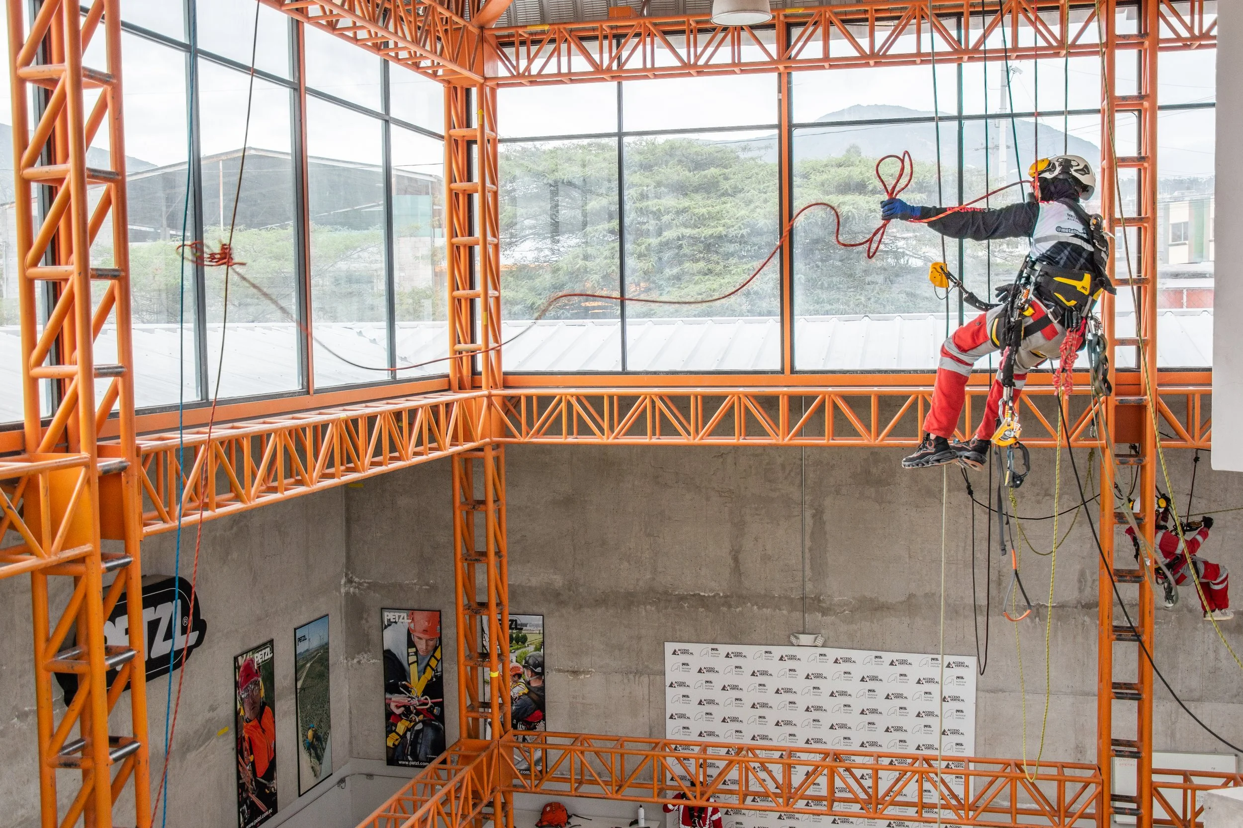 Persona en equipo de escalada en interiores, colgado en una estructura de metal naranja, realizando una actividad desafiante de altura y escalada.