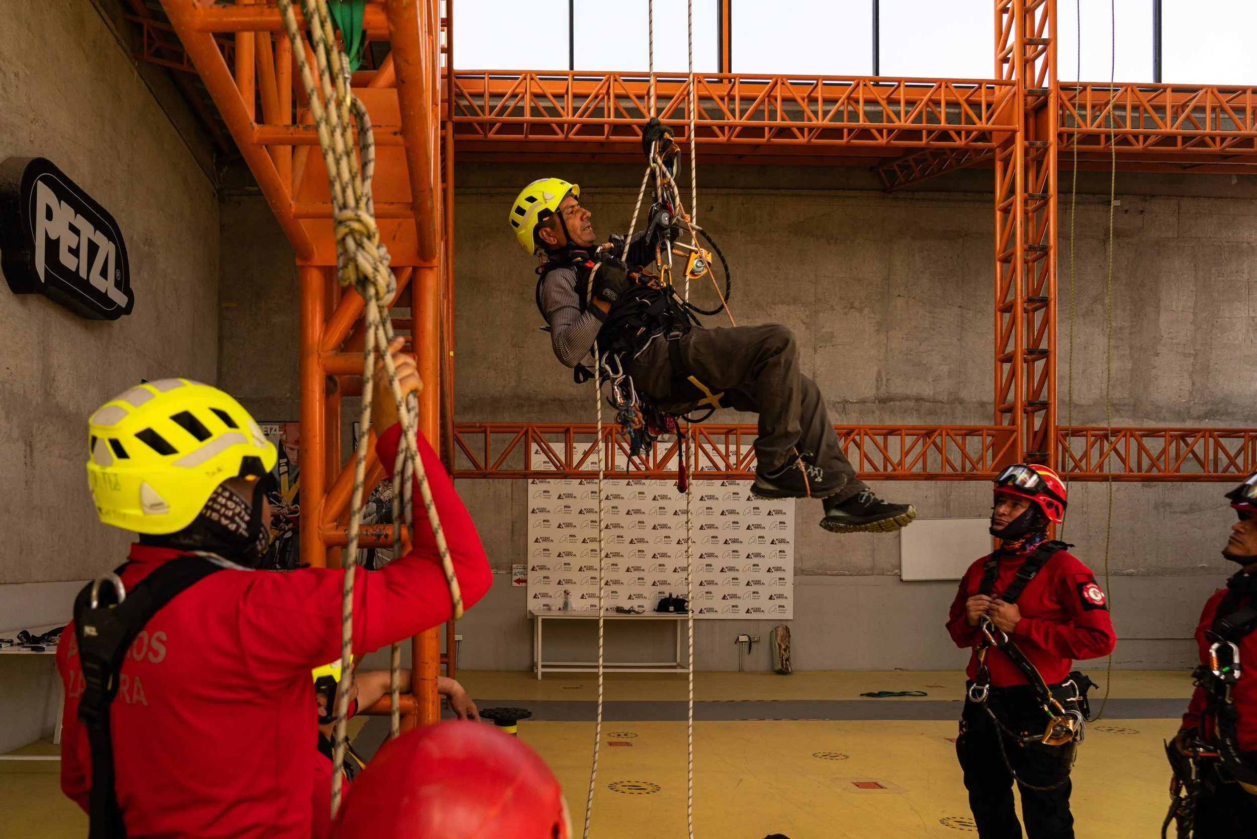 Diversas personas equipadas con cascos y arneses de seguridad participan en un entrenamiento de rescate en interiores, con un hombre usando un arnés y cuerda suspendido en el aire, en un espacio con estructura de metal naranja y paredes de cemento.