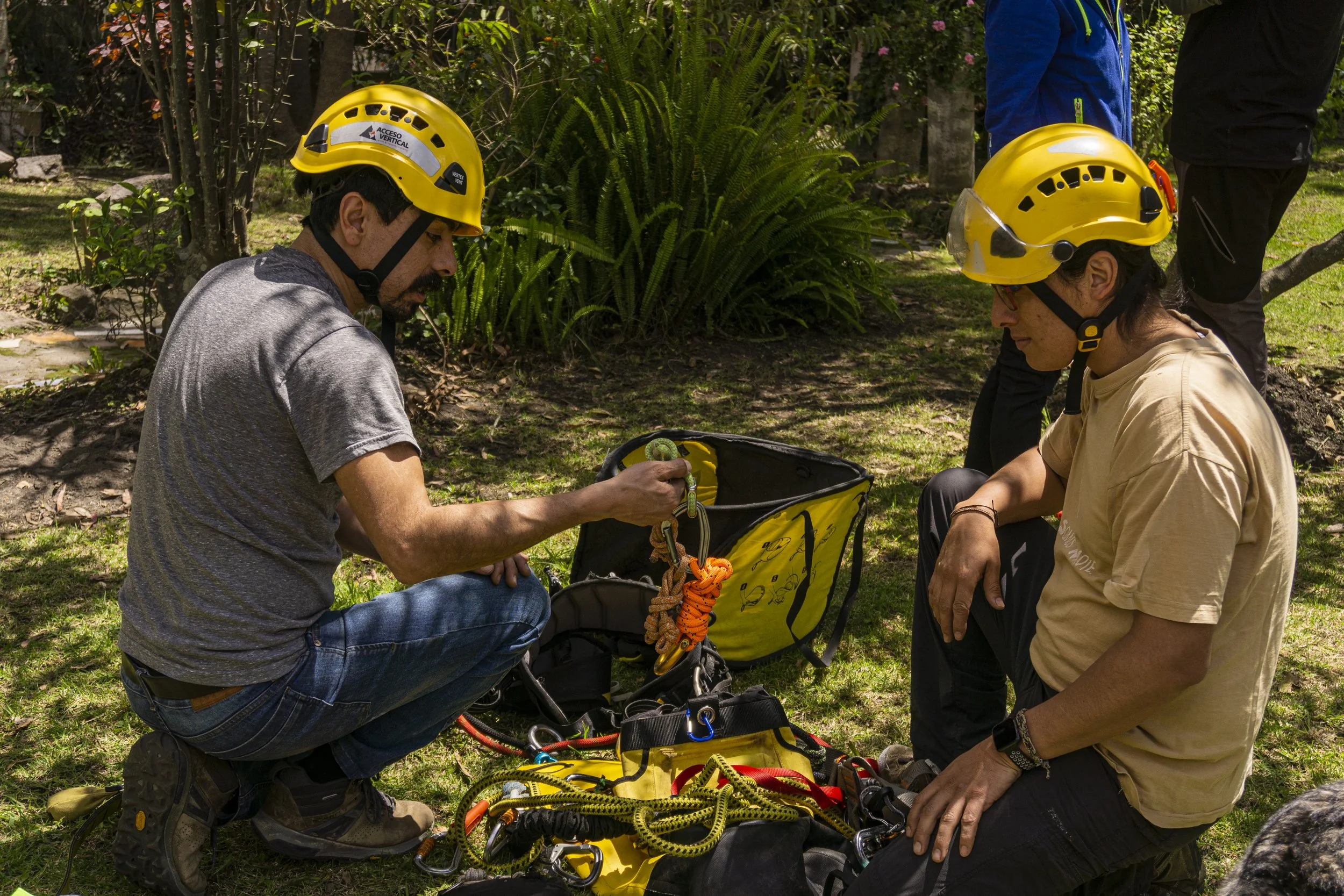 Dos personas con casco de seguridad negro y amarillo en una sesión de entrenamiento de escalada o rescate, revisando equipamiento en un parque con árboles y vegetación.