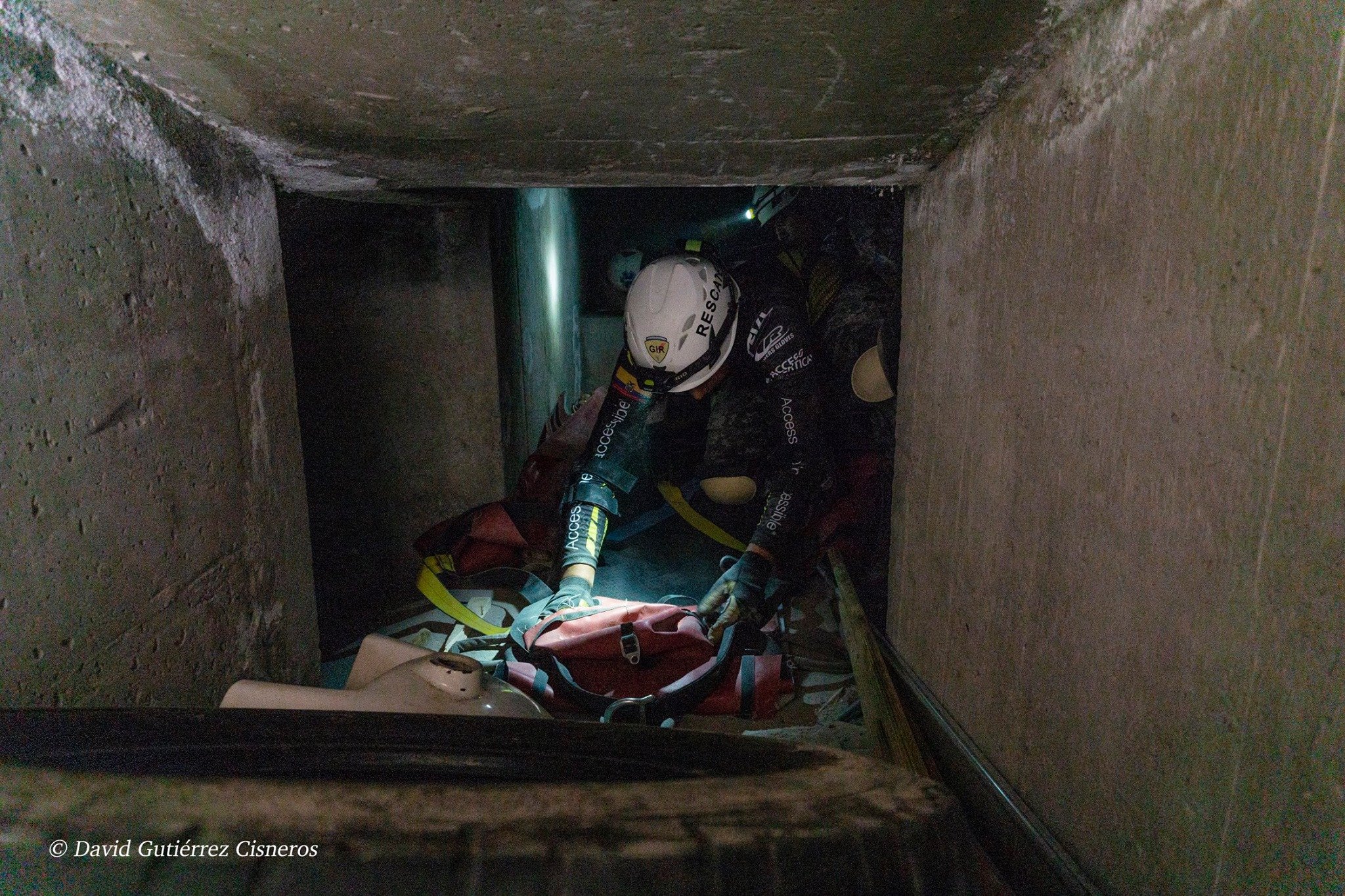 Rescatista sentado en un espacio estrecho con equipo de protección y mochila, en un entorno de pared de concreto en un ambiente de rescate vertical.
