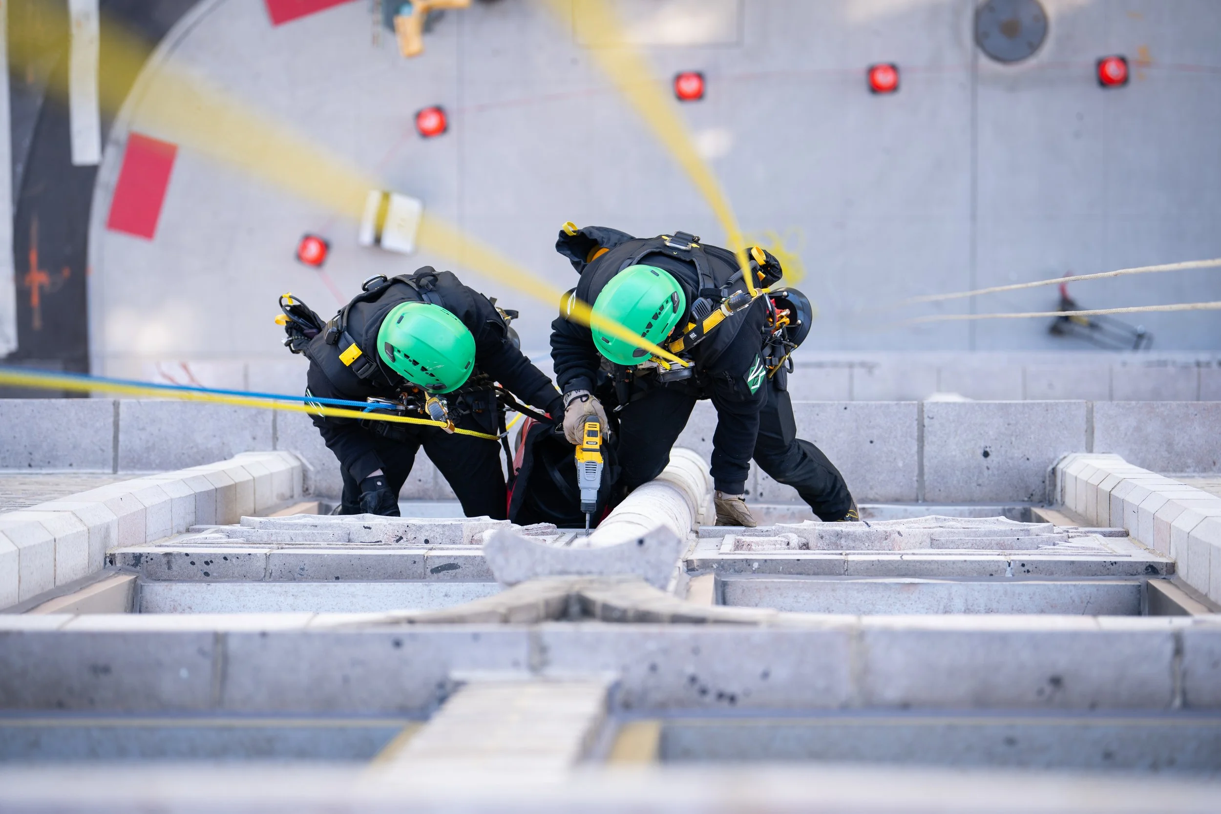 Dos rescatistas con cascos verdes y arnés trabajando en un edificio alto, asegurando una manguera grande con líneas de seguridad amarillas.