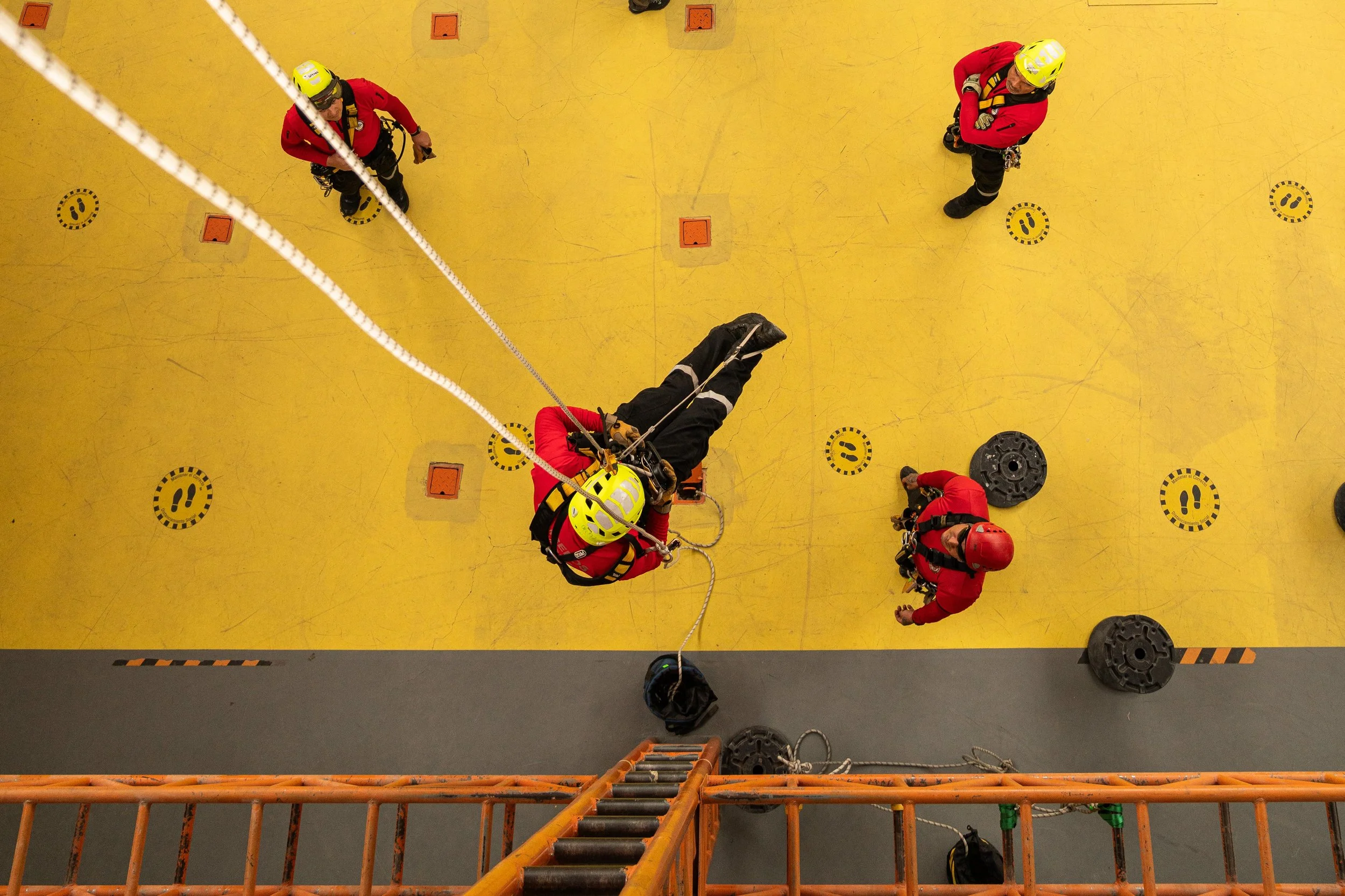 Cuatro bomberos en un entrenamiento en un edificio, con un bombero colgado de una cuerda, aprendiendo técnicas de rescate en alturas.