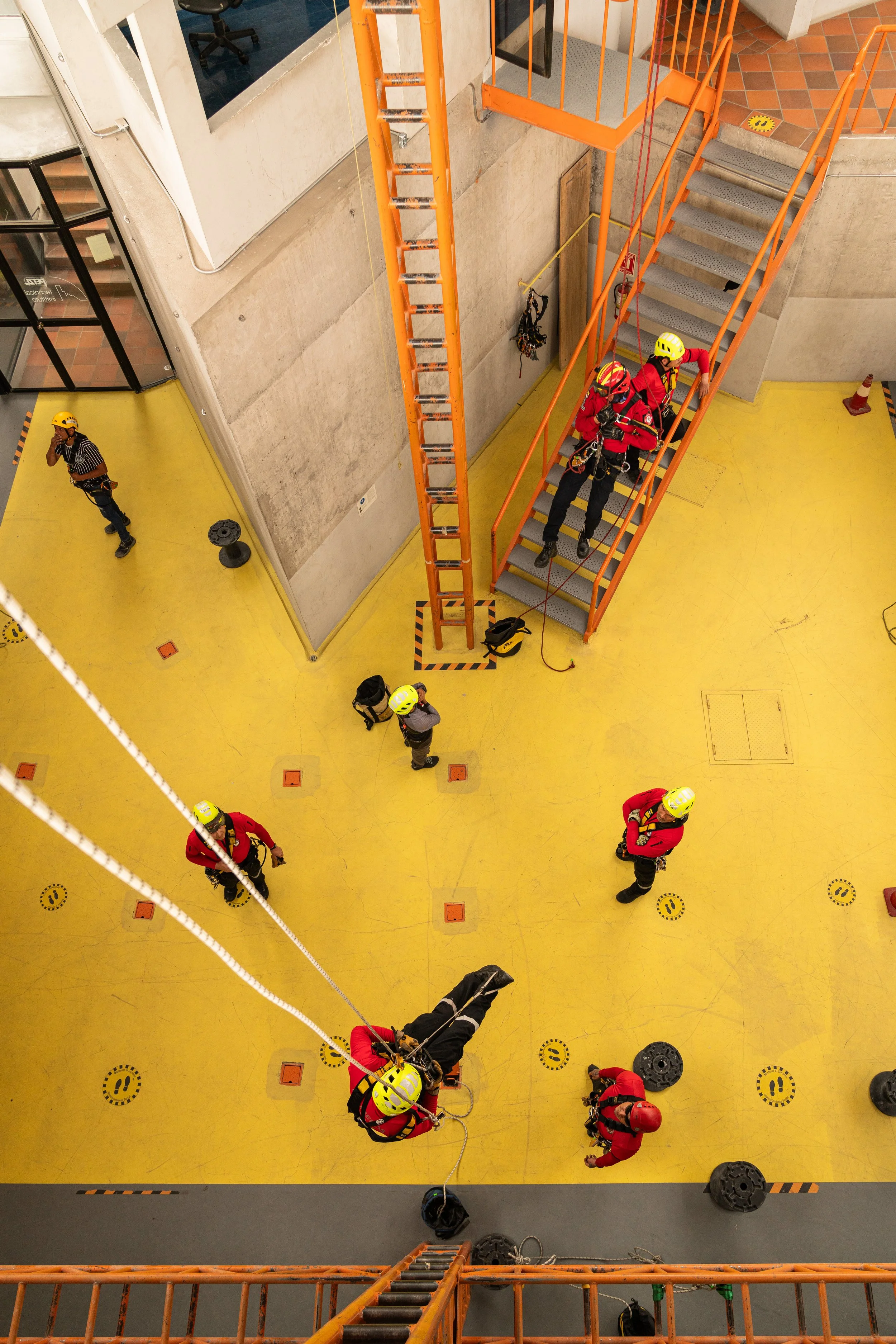 Técnicos de rescate en entrenamiento en un área industrial con suelo amarillo, escaleras y estructura de hierro naranja, varios miembros en cascos y ropa de seguridad participan en una simulación de rescate.