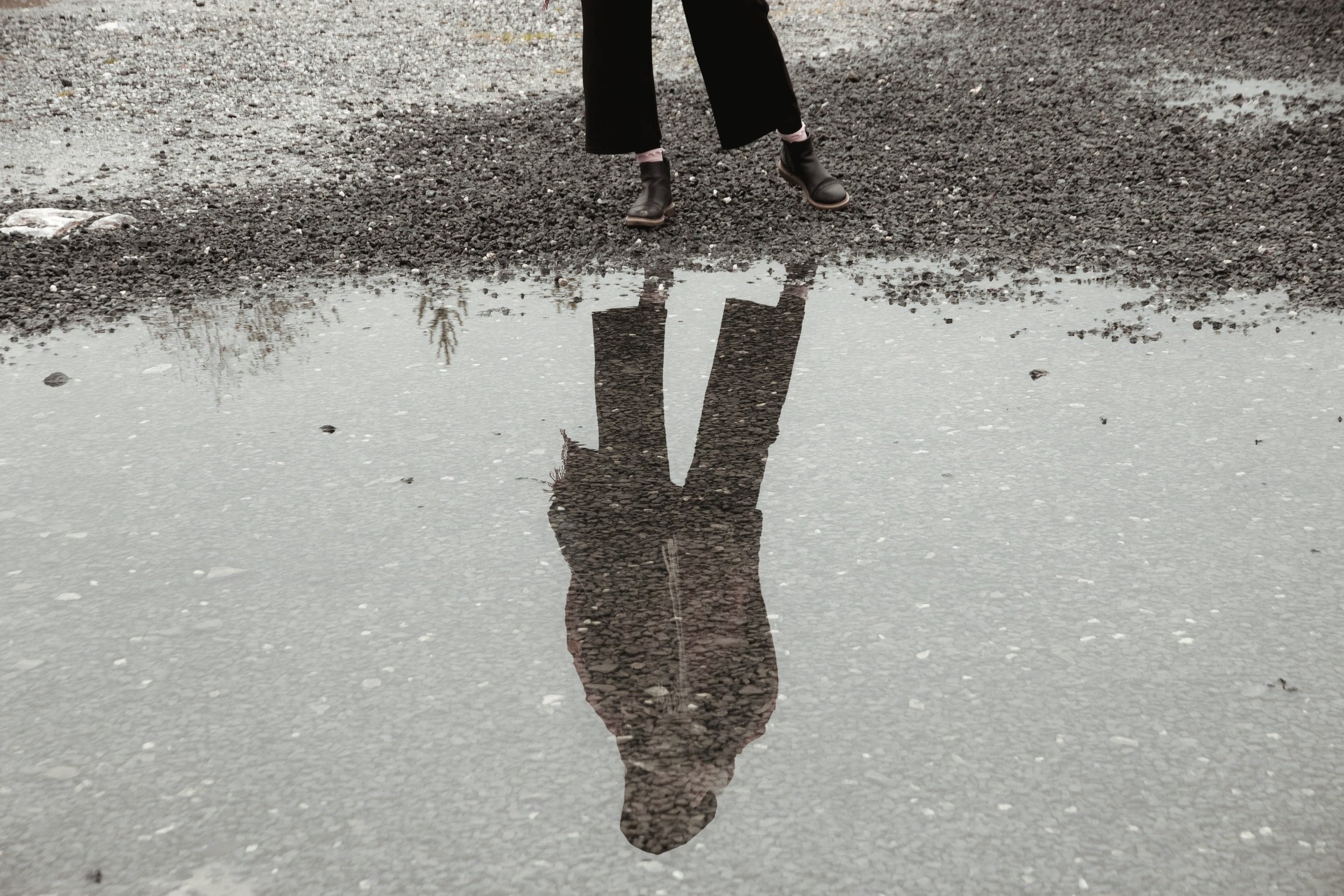 Reflection of a person standing near a puddle on a gravel ground, showing only their legs and shoes.