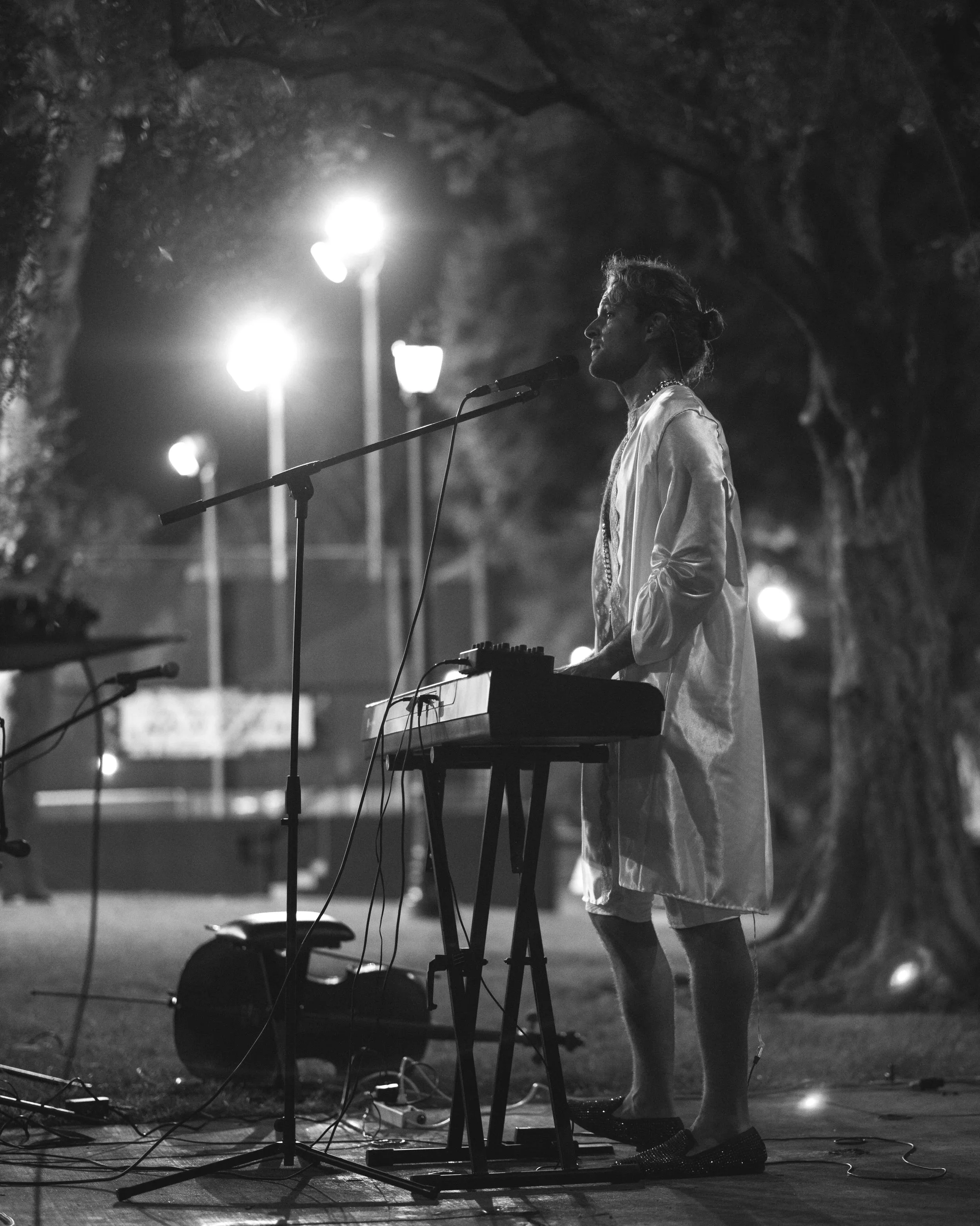 Natti Vogel performing music outdoors at night, standing at a keyboard with a microphone, illuminated by streetlights. For WEHO Arts in California.