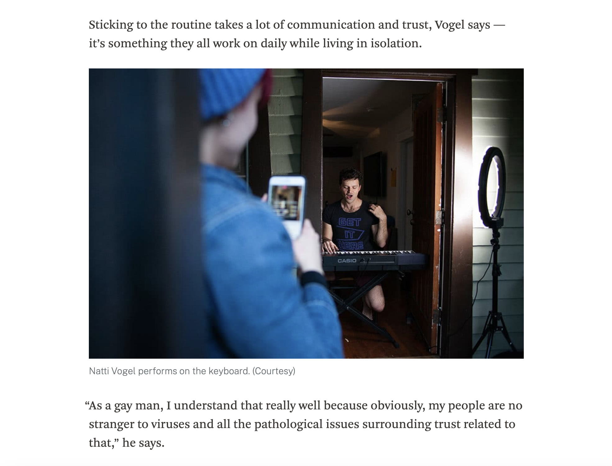 A person in the foreground is taking a photo or video with their smartphone of a man performing on a keyboard inside a room. Natti Vogel is wearing a black t-shirt with blue text and is singing.