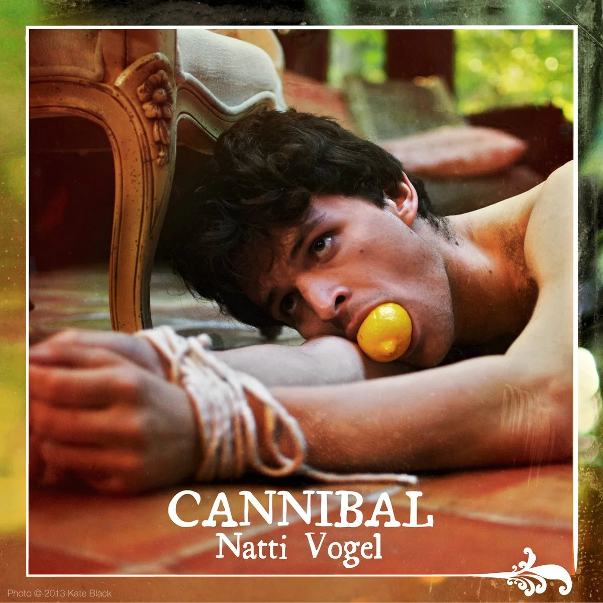 A man (NATTI VOGEL) lying on the floor with a lemon in his mouth, resting his head on a vintage wooden piece of furniture, in a room with green foliage outside the window, with the text 'CANNIBAL Natti Vogel' at the bottom.