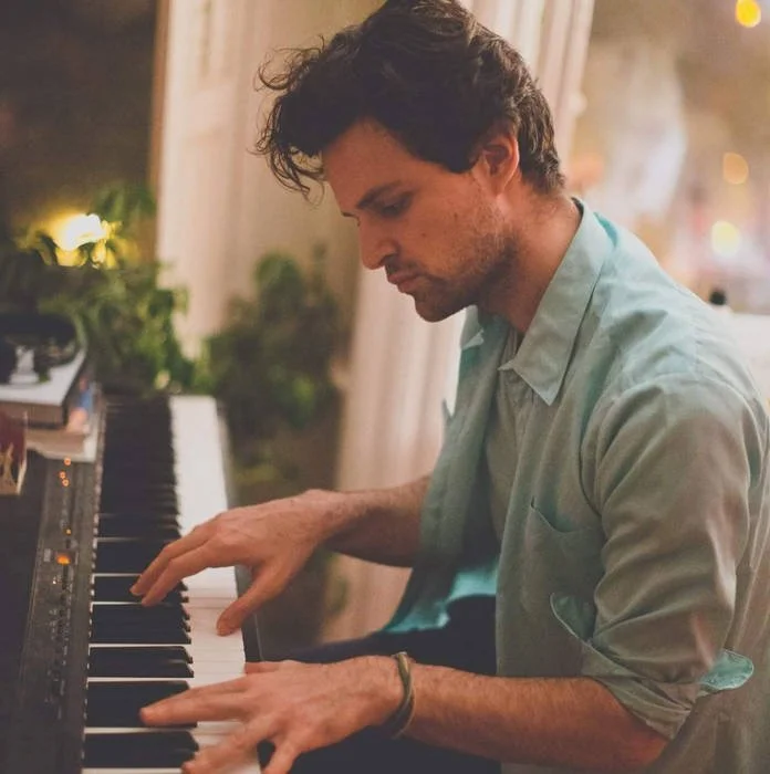 Natti Vogel playing a piano indoors with warm lighting and plants in the background.